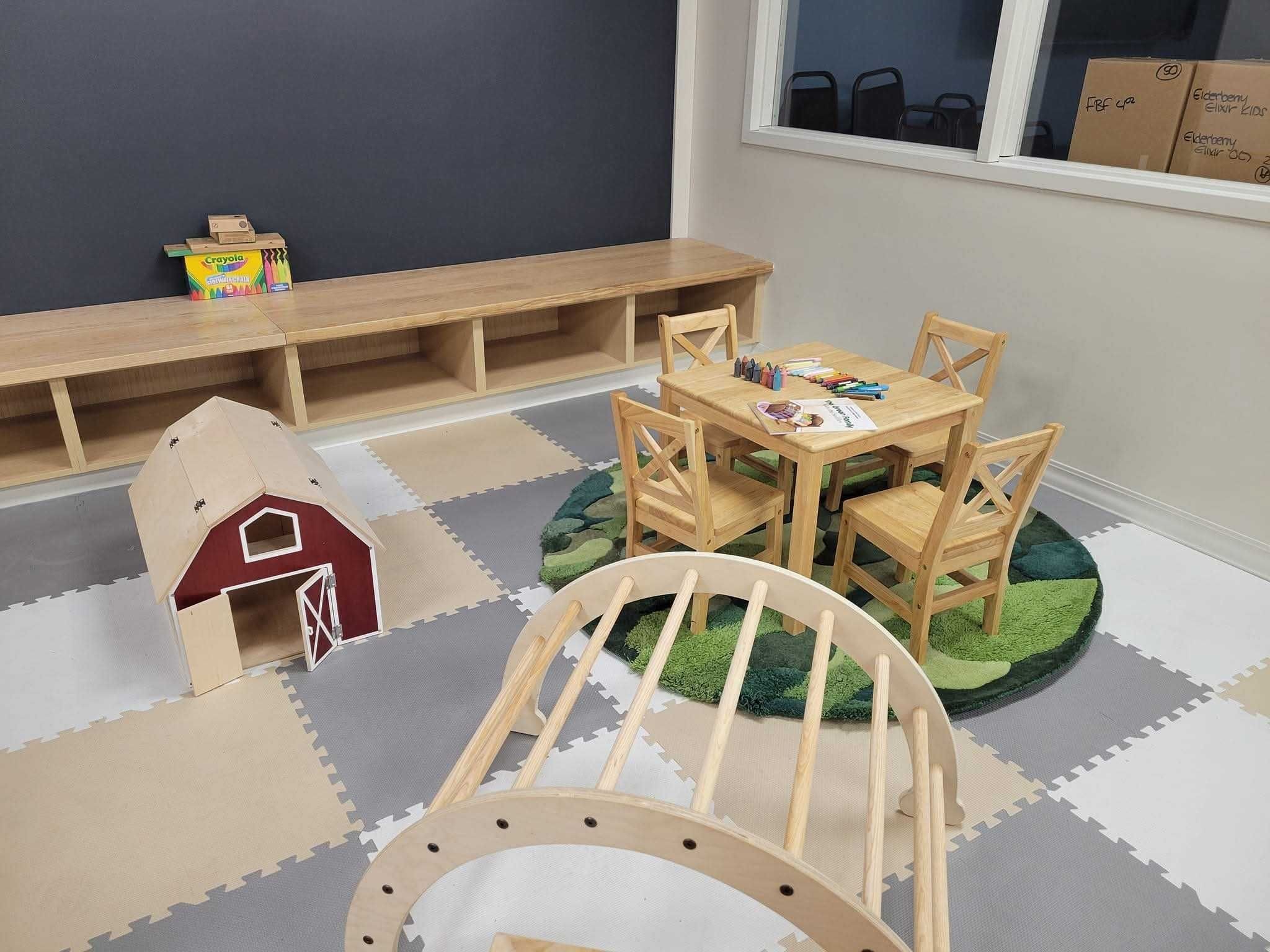 Kids play area in Earthley retail store featuring wooden table with chairs on green grass rug, wooden climbing rocker, red barn playhouse, Crayola crayons, and interlocking foam floor mats