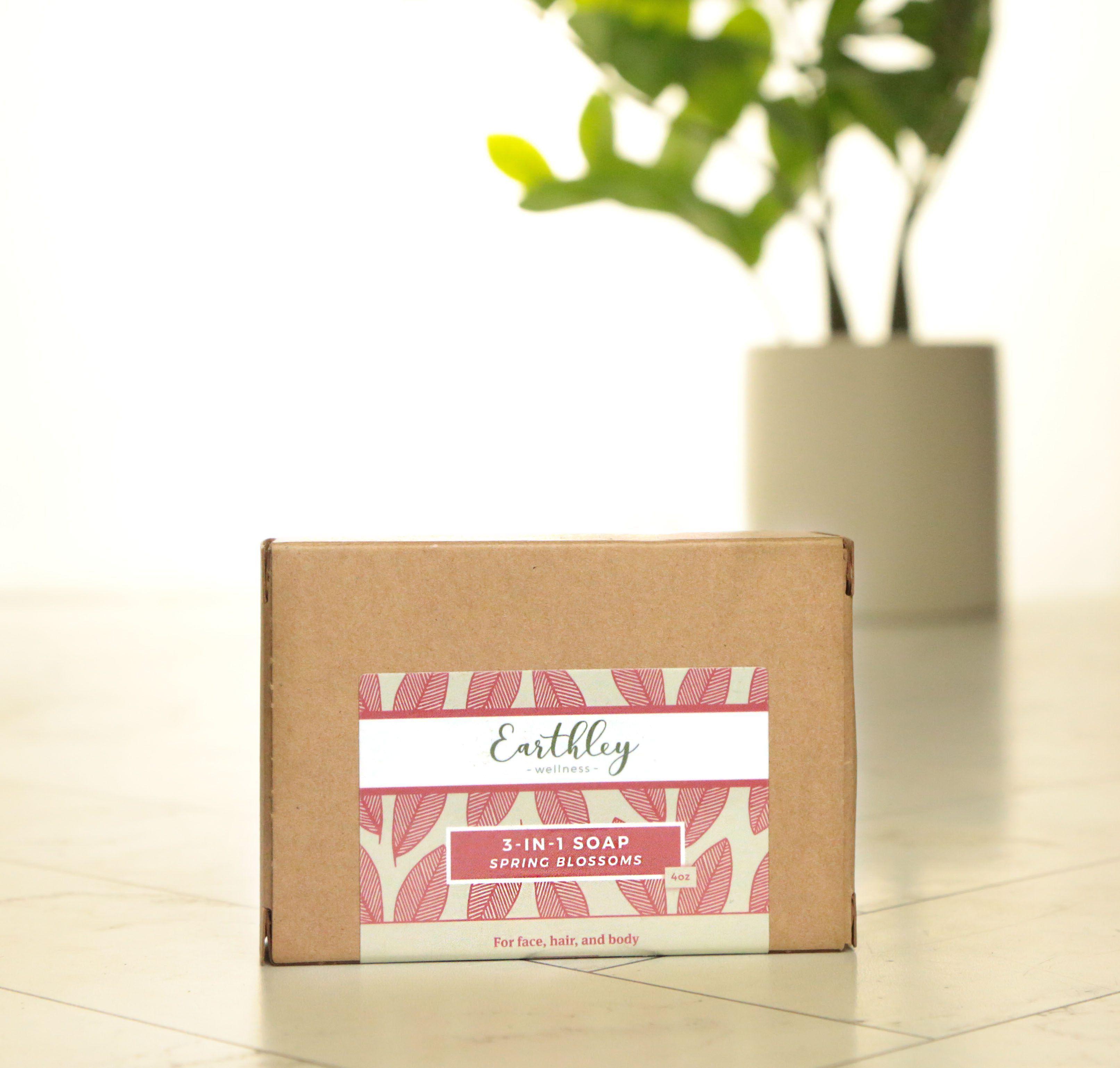 Earthley 3-in-1 Soap Spring Blossoms 4oz tan kraft box with pink coral leaf pattern label on white marble tile with green plant in cream ceramic pot