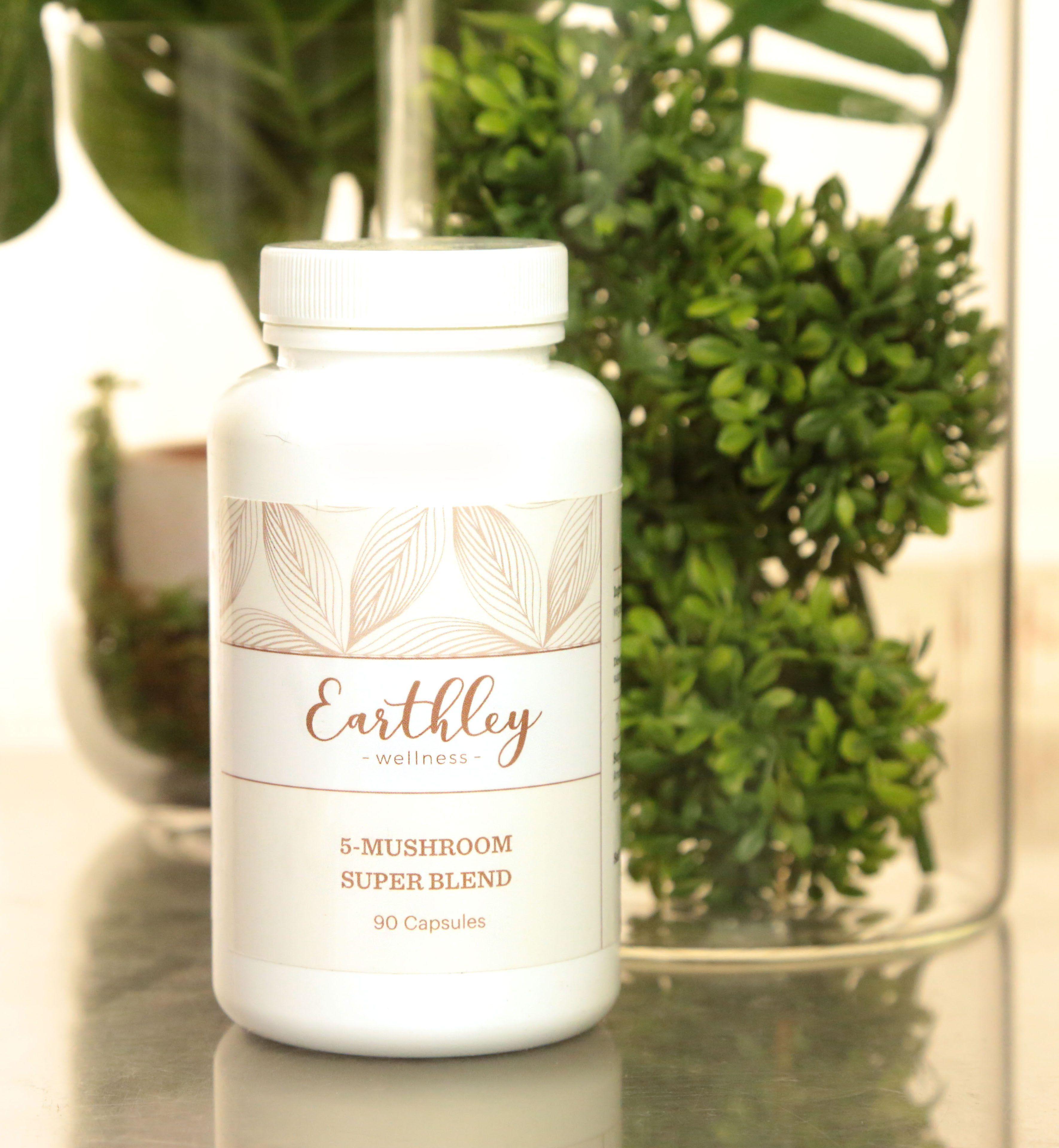 Earthley 5-Mushroom Super Blend 90 capsules white plastic bottle with white lid and tan leaf pattern label on gray marble surface with green boxwood and tropical plants in clear glass terrarium