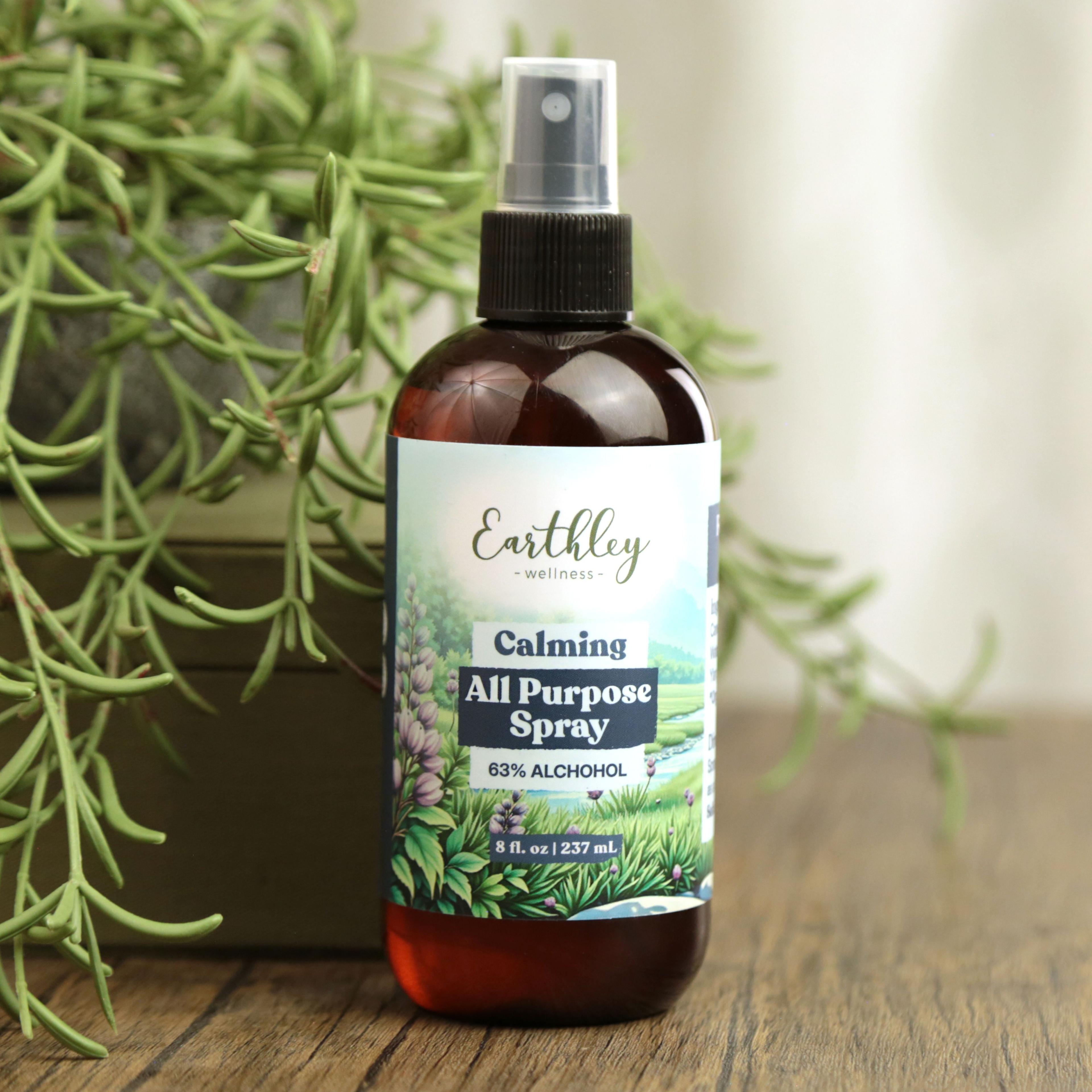Earthley Calming All Purpose Spray 8 oz amber spray bottle with scenic meadow and lavender field label showing 63% Alcohol content, on wooden surface with trailing succulent plant