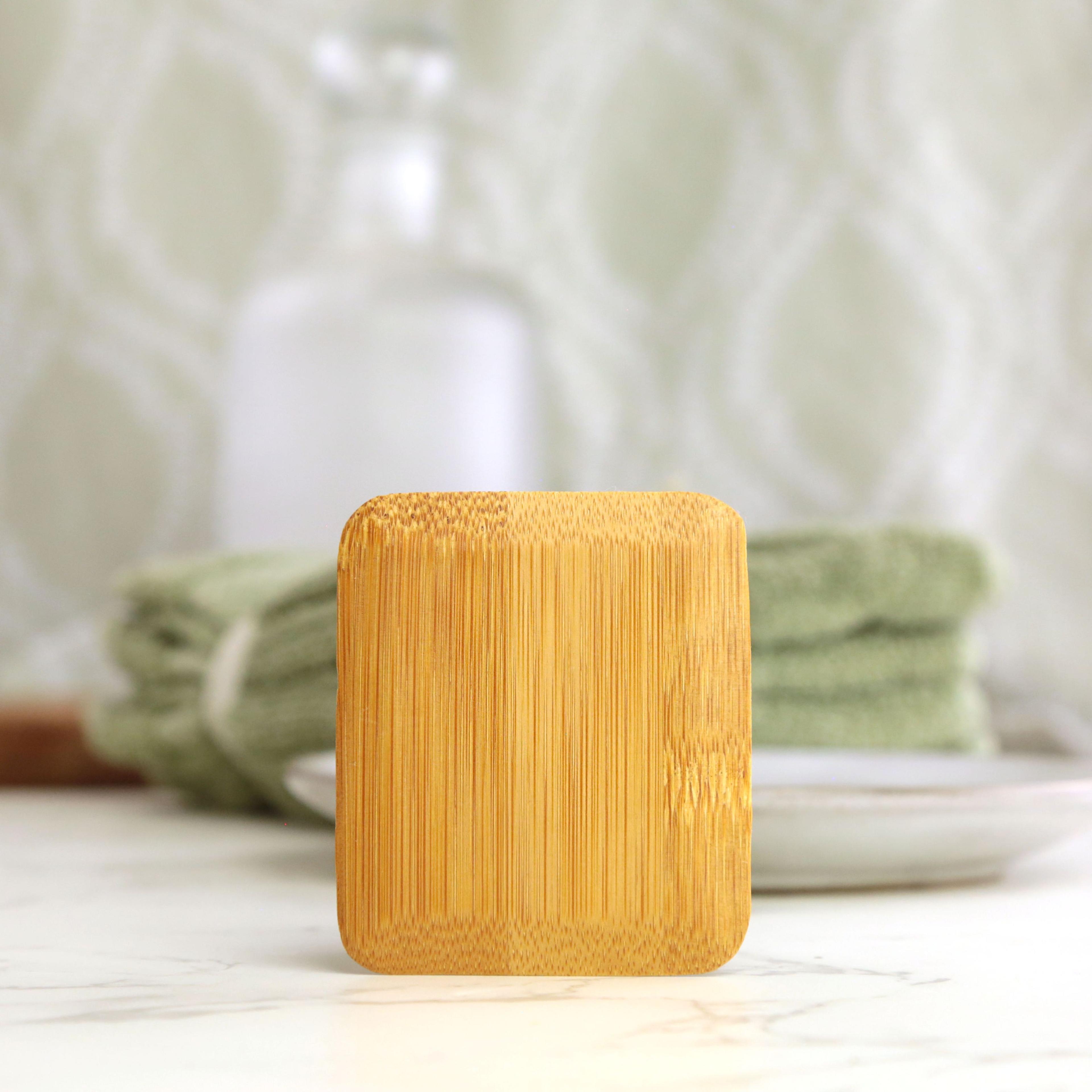 Bamboo dish scraper front view showing natural tan golden bamboo square pad on white marble countertop with sage green towel on white plate and blurred glass bottle