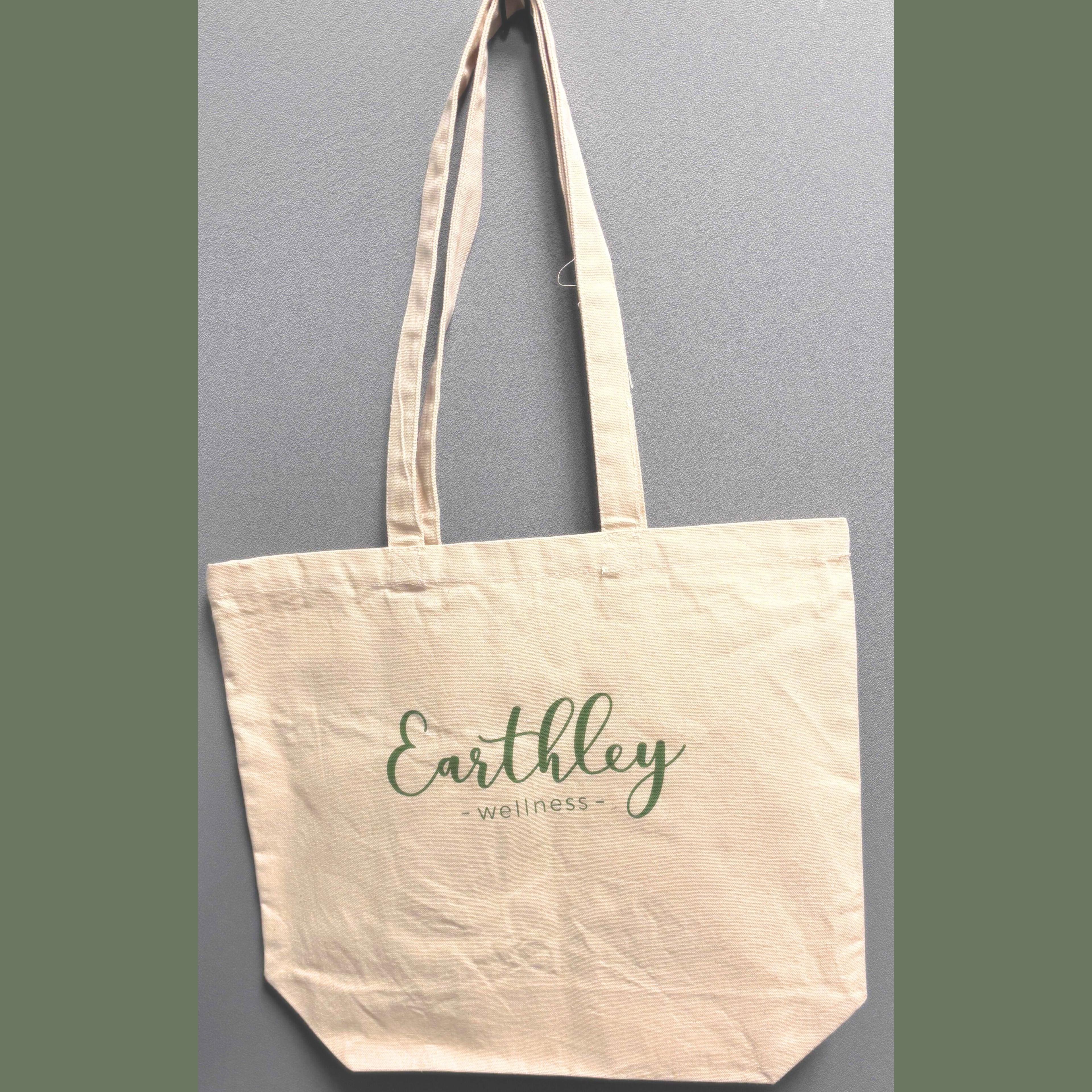 Natural canvas tote bag with Earthley Wellness logo in green script hanging against gray background