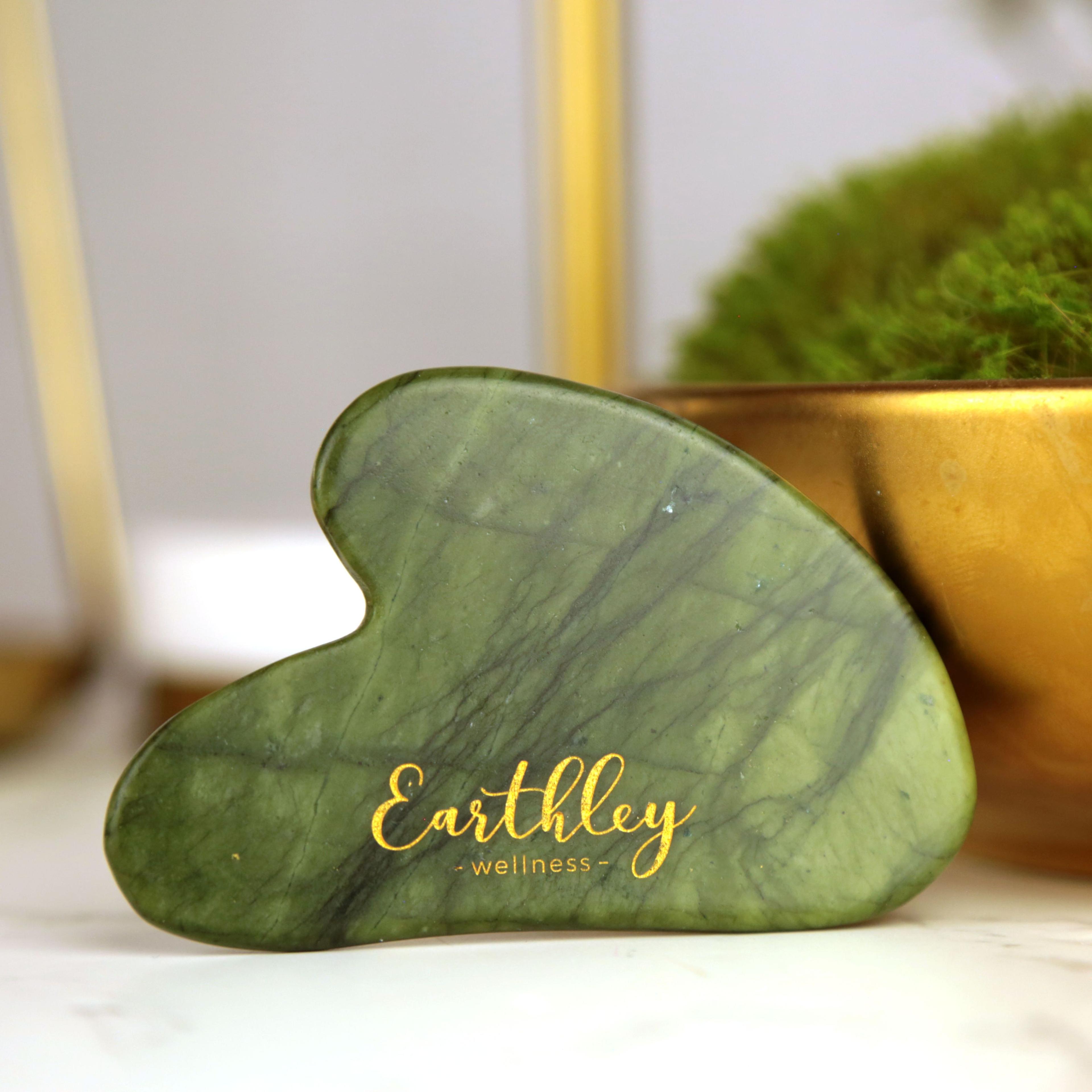 Green jade gua sha tool with Earthley Wellness logo in gold lettering, brass bowl with herbs in background