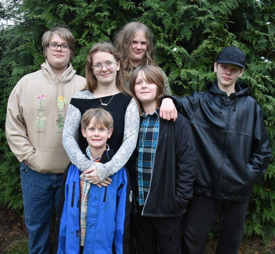 Group of six children and young adults posing together outdoors in front of evergreen bushes, wearing casual clothing including hoodies and jackets