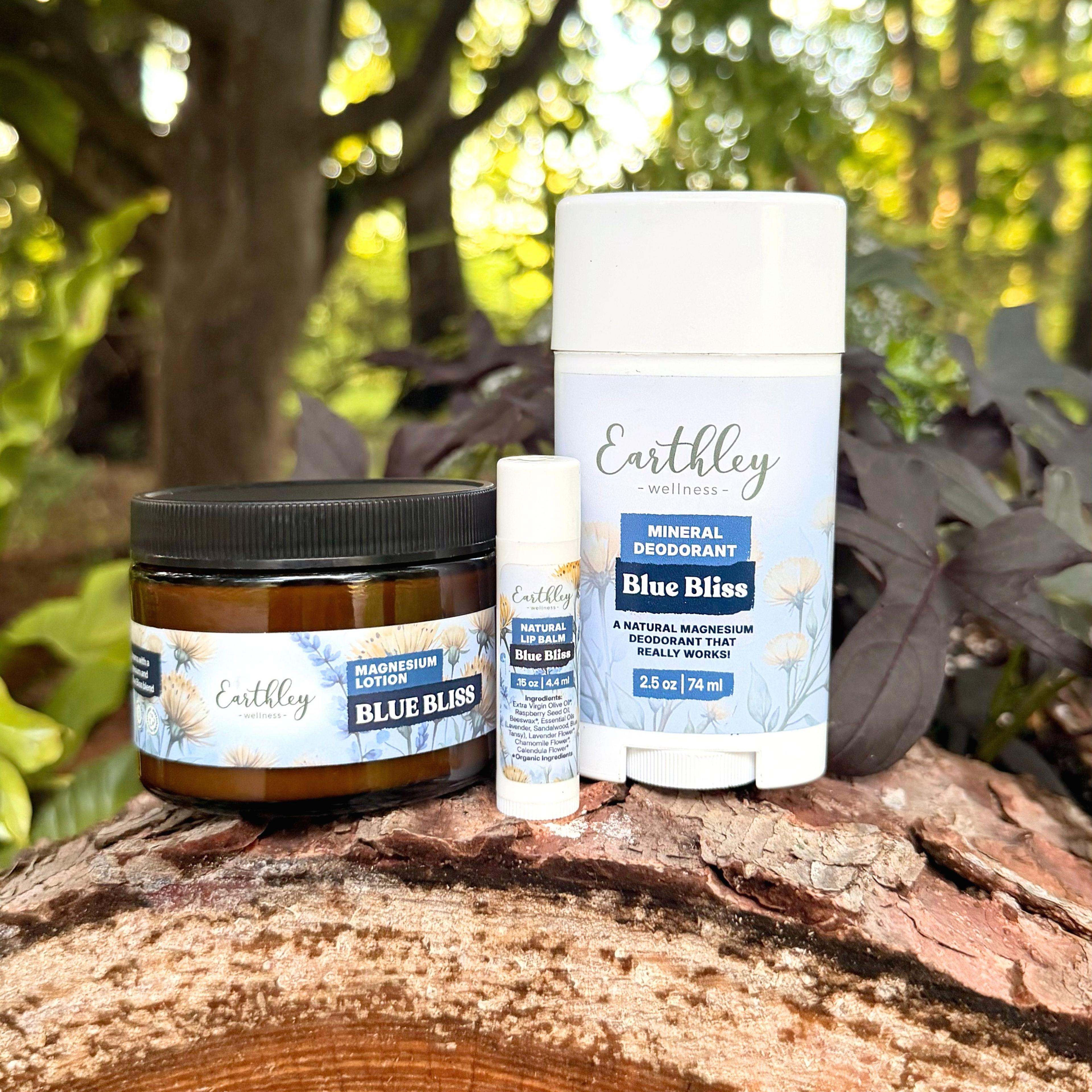 Earthley Wellness Blue Bliss trio: magnesium lotion jar, lip balm, and mineral deodorant stick on log outdoors.