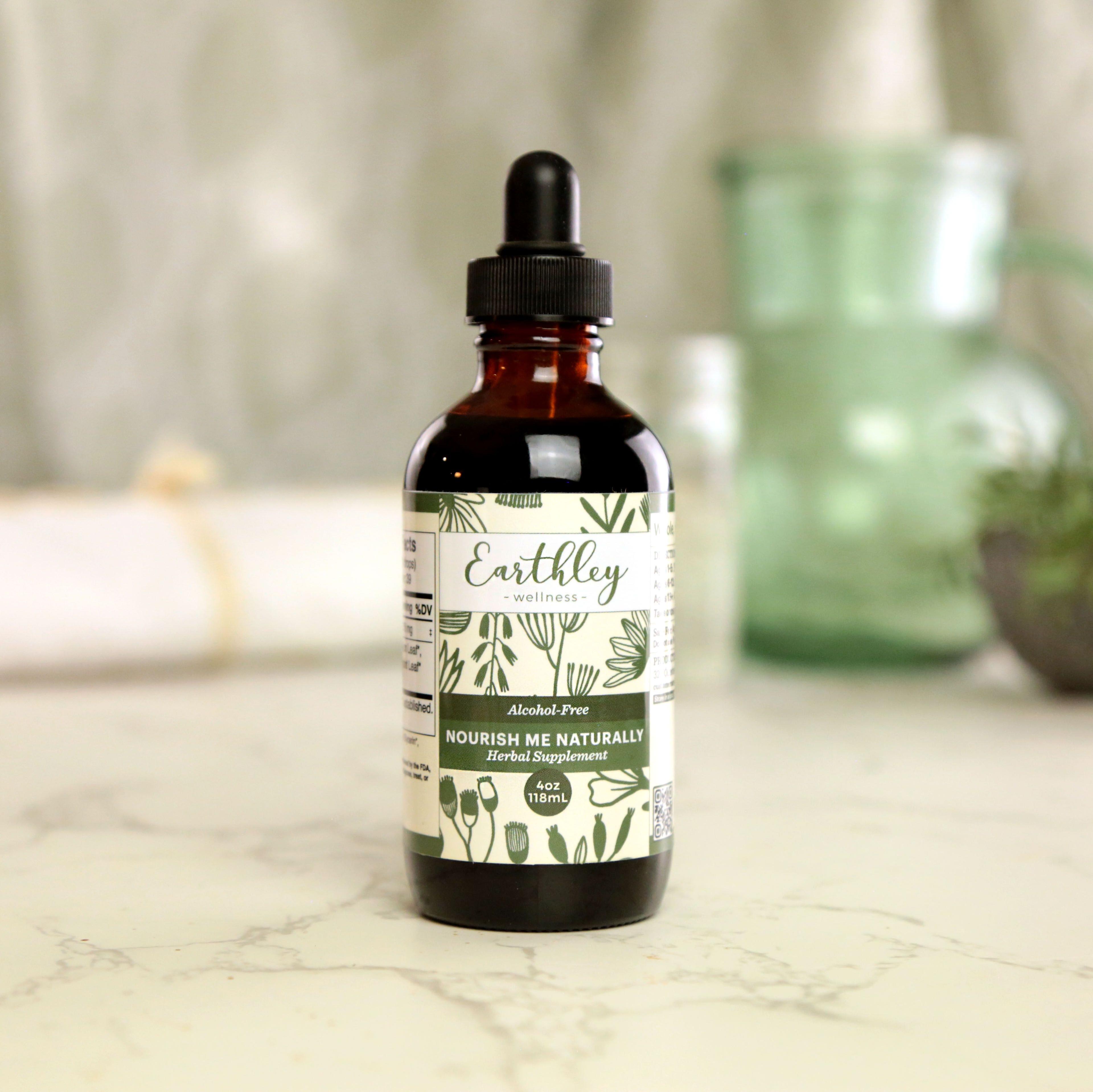 Earthley Nourish Me Naturally 4oz 118ml amber glass dropper bottle with black cap and sage green botanical floral label on white marble with green glass pitcher