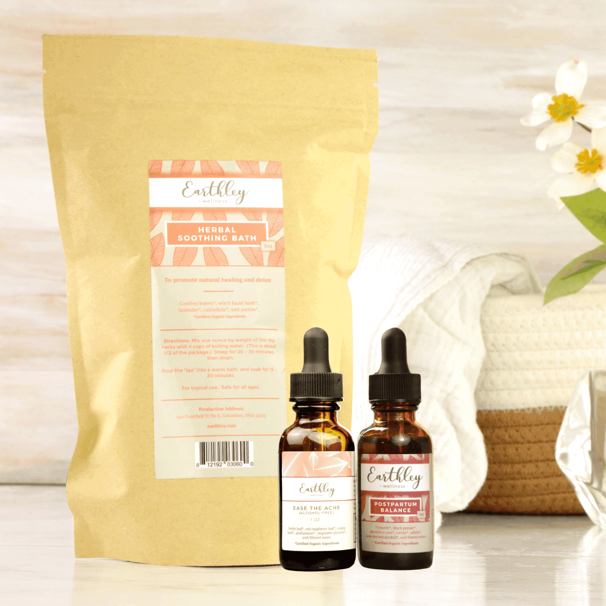 Earthley Postpartum Mama bundle showing Herbal Soothing Bath bag, Ease the Ache tincture, and Postpartum Balance tincture with white towels and flowers