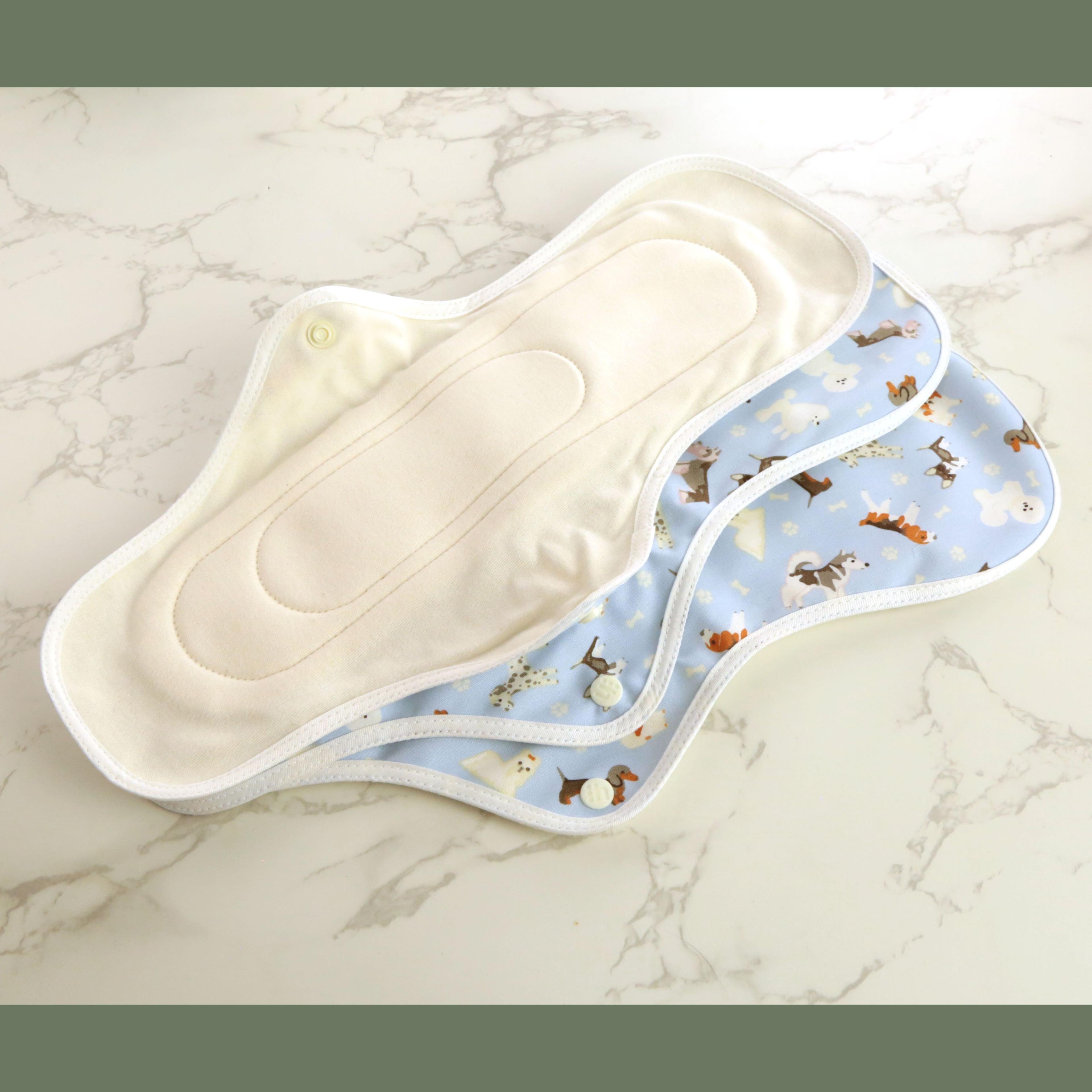 Three hannahPAD reusable sanitary pads in large overnight size showing cream organic cotton interior lining and cute dog pattern on light blue fabric exterior with snap closures, arranged fanned out on marble surface