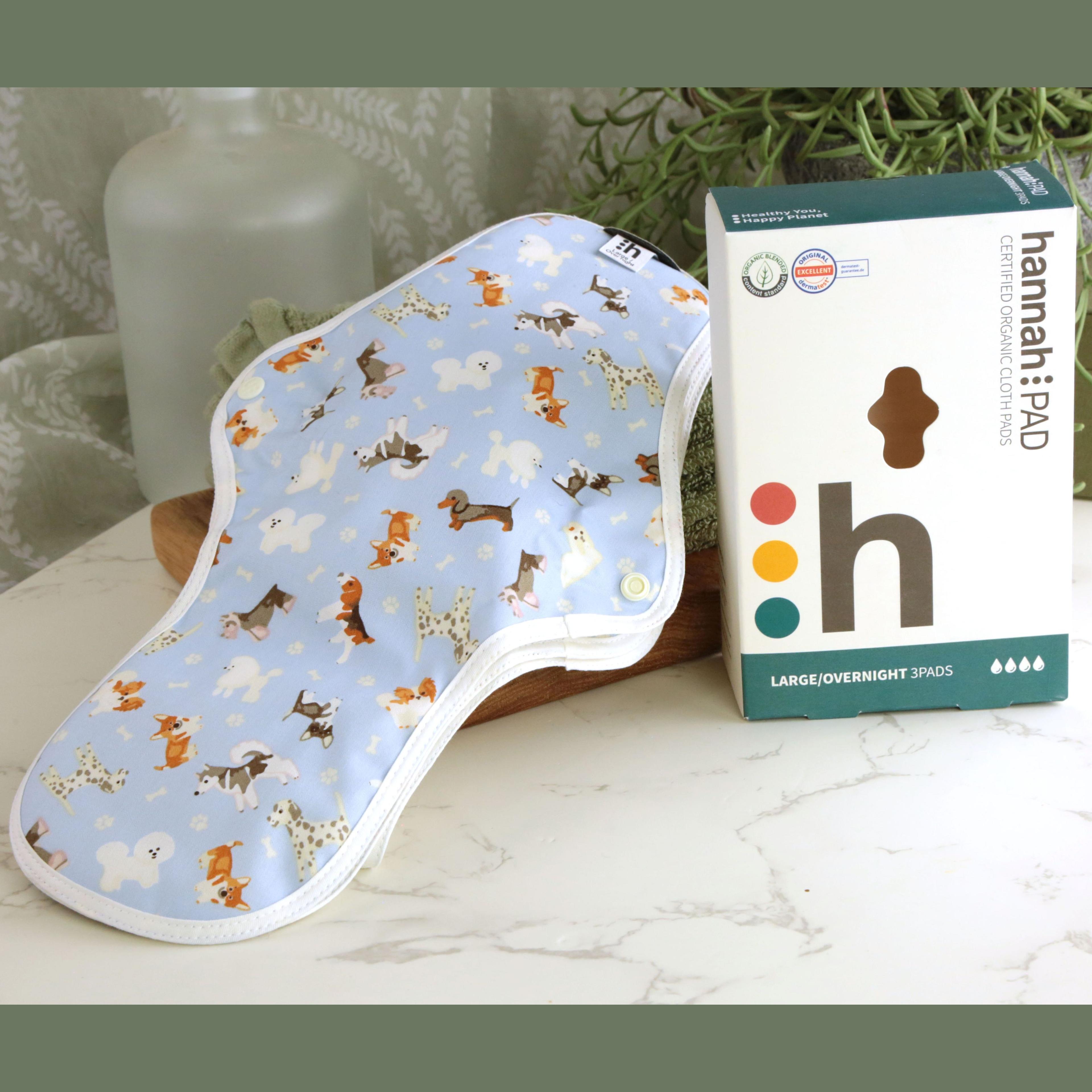 hannahPAD Certified Organic Cloth Pads Large Overnight 3 Pads set with cute dog pattern on light blue fabric, displayed with teal and white branded box, on marble surface with green towels and trailing succulent plant in background