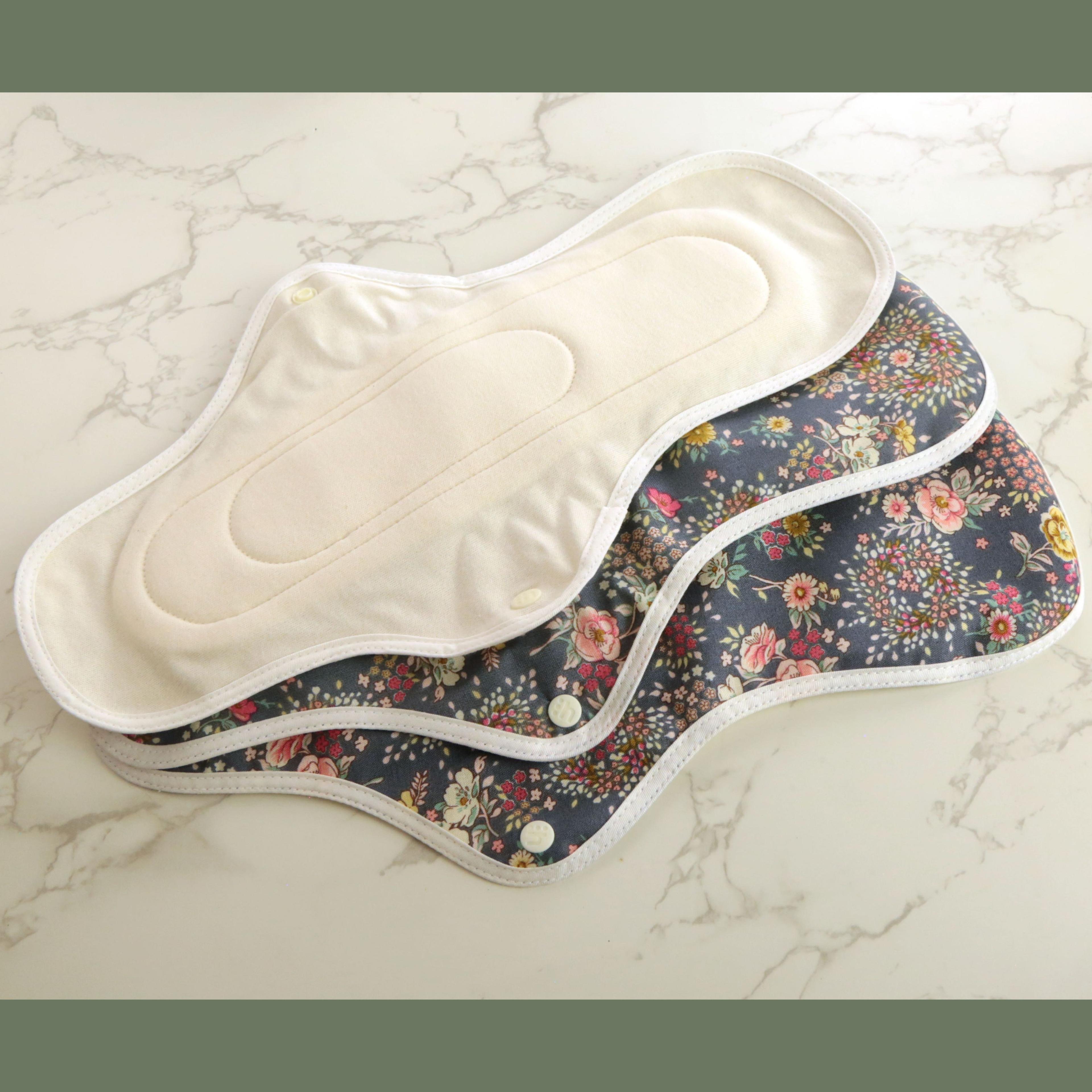 Three hannahPAD reusable sanitary pads in large overnight size showing cream organic cotton interior lining and colorful floral pattern on dark gray fabric exterior with snap closures, arranged fanned out on marble surface
