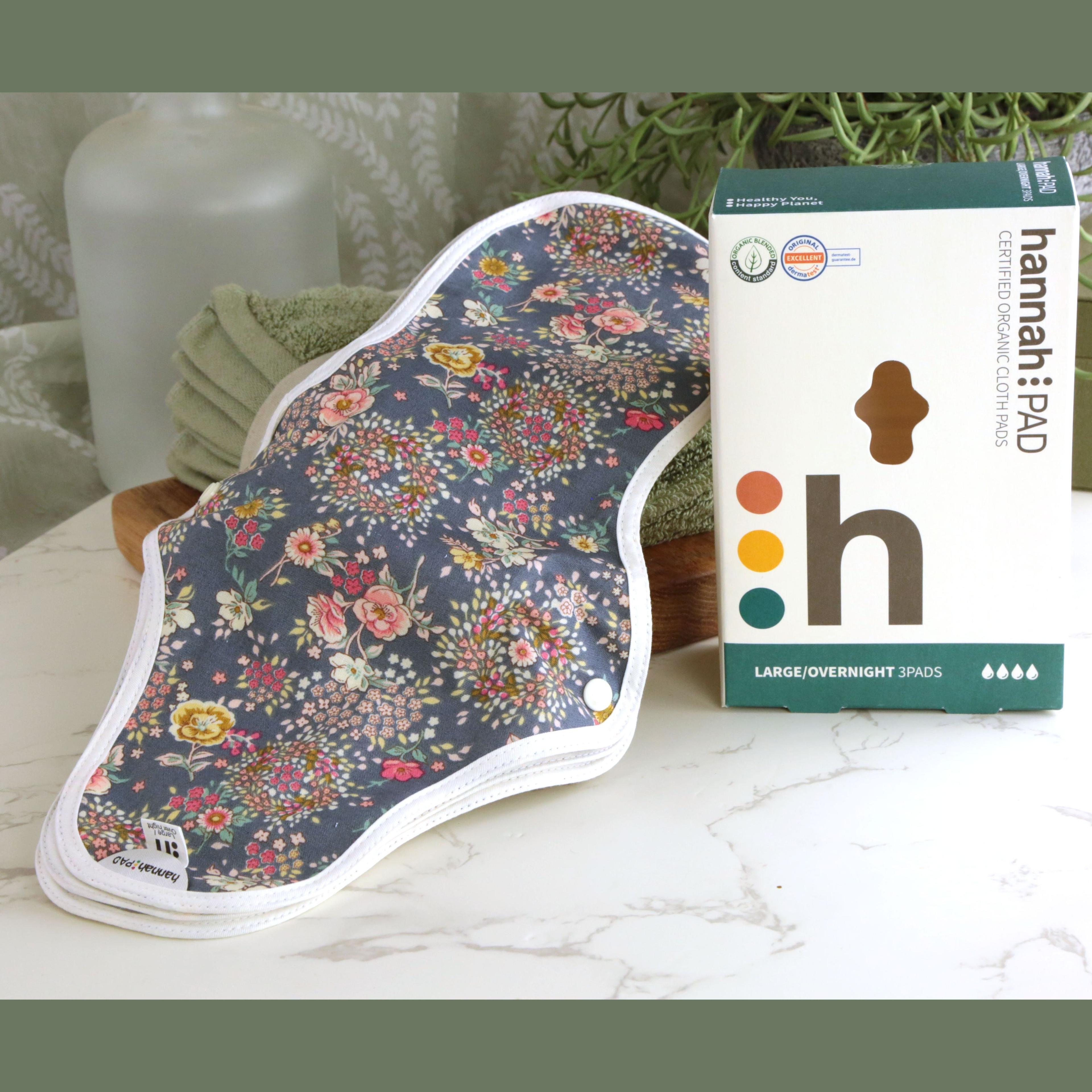 hannahPAD Certified Organic Cloth Pads Large Overnight 3 Pads set with colorful floral pattern on dark gray fabric, displayed with teal and white branded box, on marble surface with green towels and trailing succulent plant in background