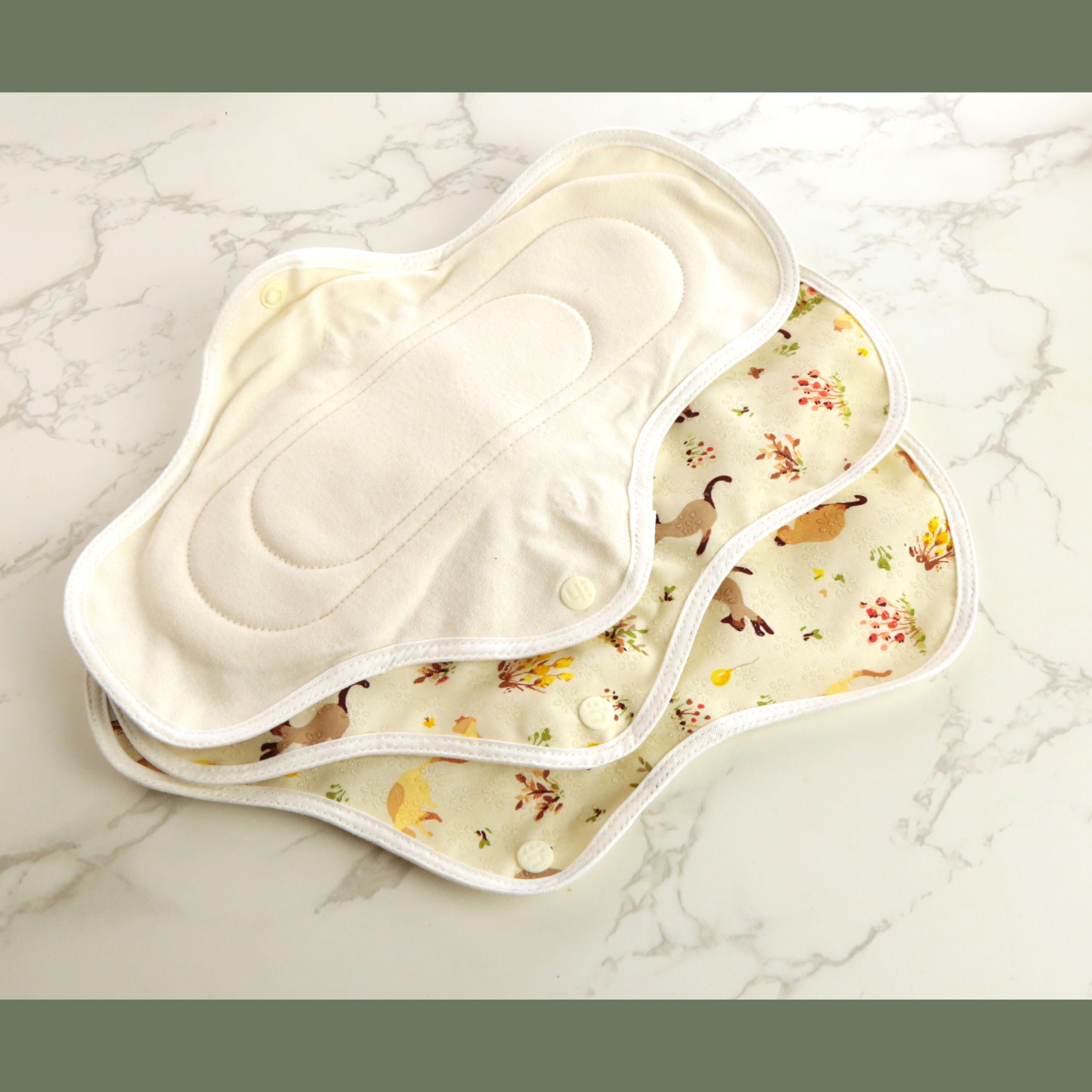 Three hannahPAD reusable sanitary pads in medium size showing cream organic cotton interior lining and cat with autumn floral pattern on cream fabric exterior with snap closures, arranged fanned out on marble surface