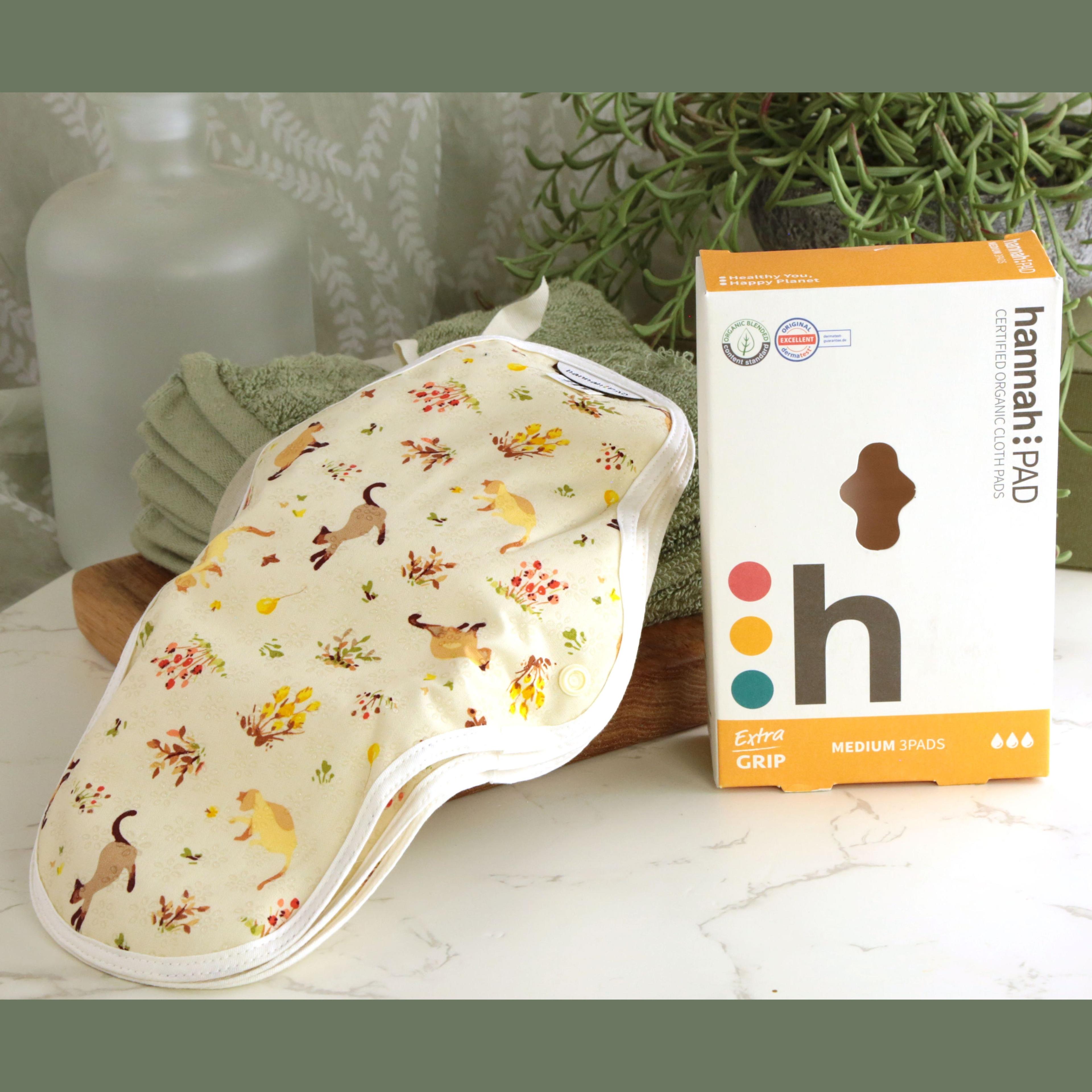 hannahPAD Certified Organic Cloth Pads Medium 3 Pads set with cat and floral pattern on cream fabric, displayed with orange and white branded box showing Extra Grip feature, on marble surface with green towels and trailing succulent plant in background