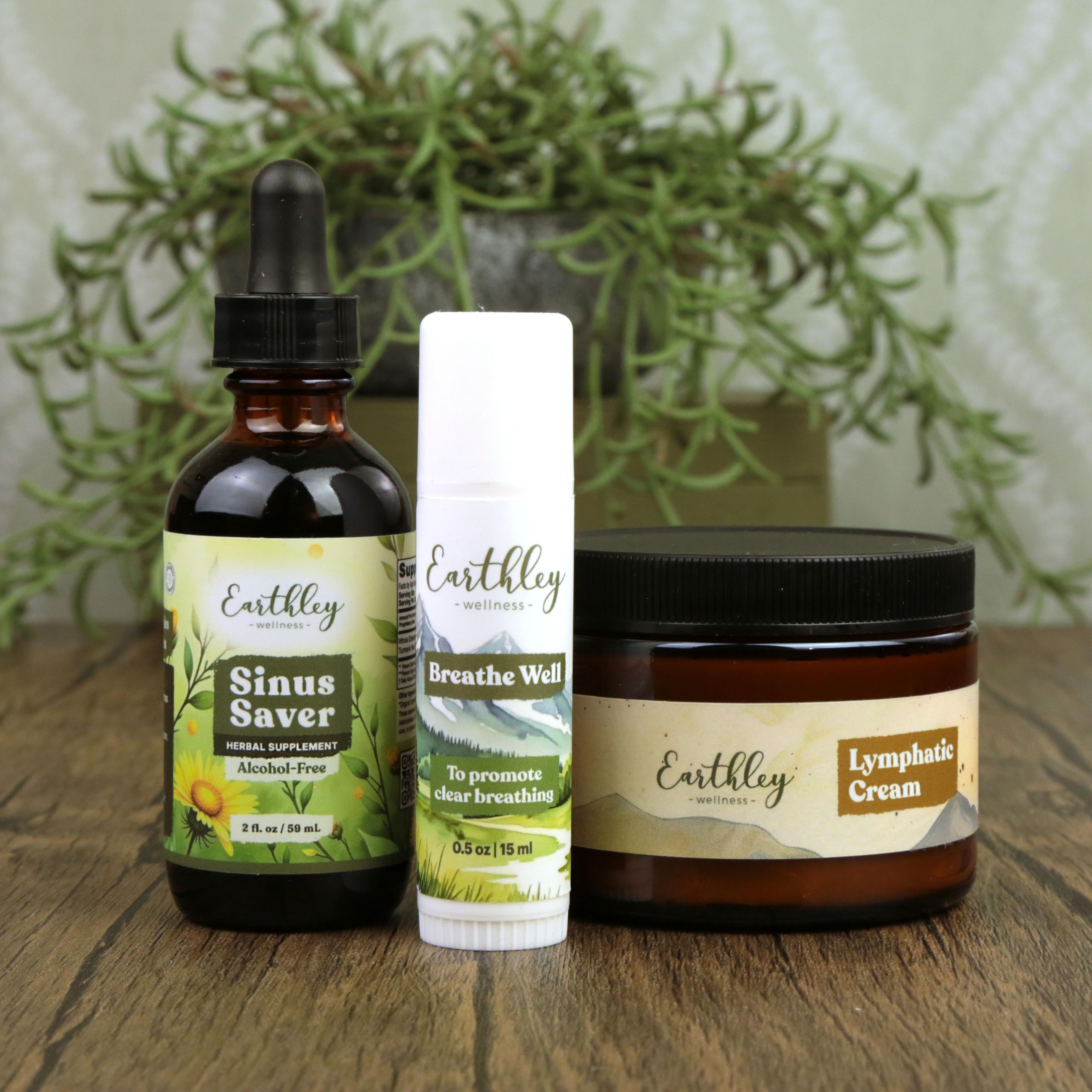 Three Earthley Wellness products on wood surface: Sinus Saver tincture, Breathe Well stick, and Lymphatic Cream jar with green plant backdrop