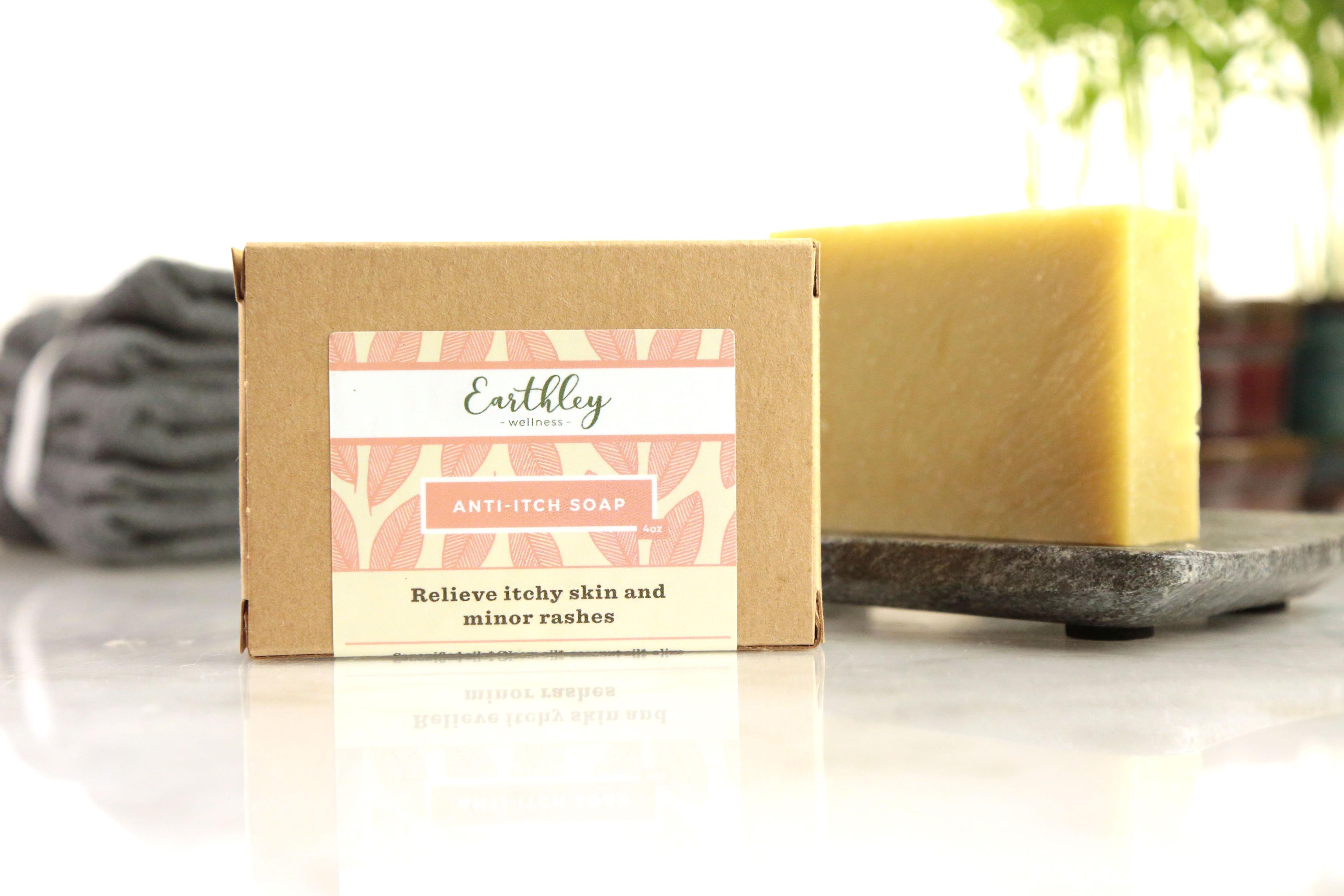 Anti-Itch Soap product photo showing tan kraft cardboard box with pink coral leaf pattern label and pale yellow soap bar on gray slate stone dish with gray towel on white marble surface and green plant in background