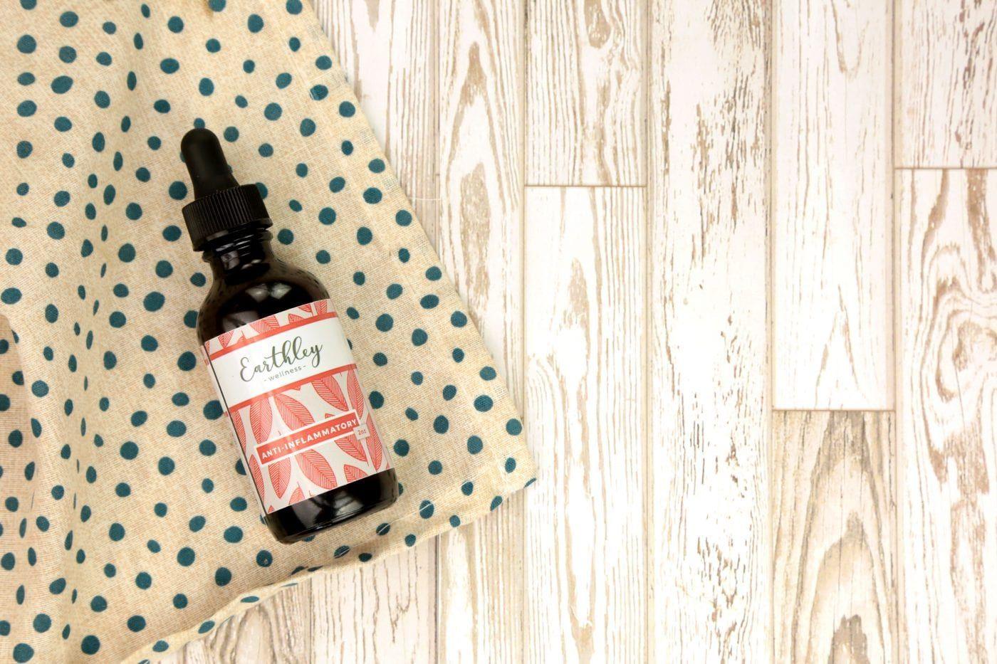 Earthley Anti-Inflammatory amber glass dropper bottle with black cap and coral pink leaf pattern label on cream fabric with teal blue polka dots on white rustic wooden surface