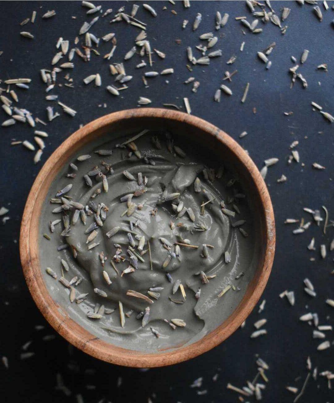 Gray clay face mask with dried lavender buds in wooden bowl, scattered lavender on dark surface