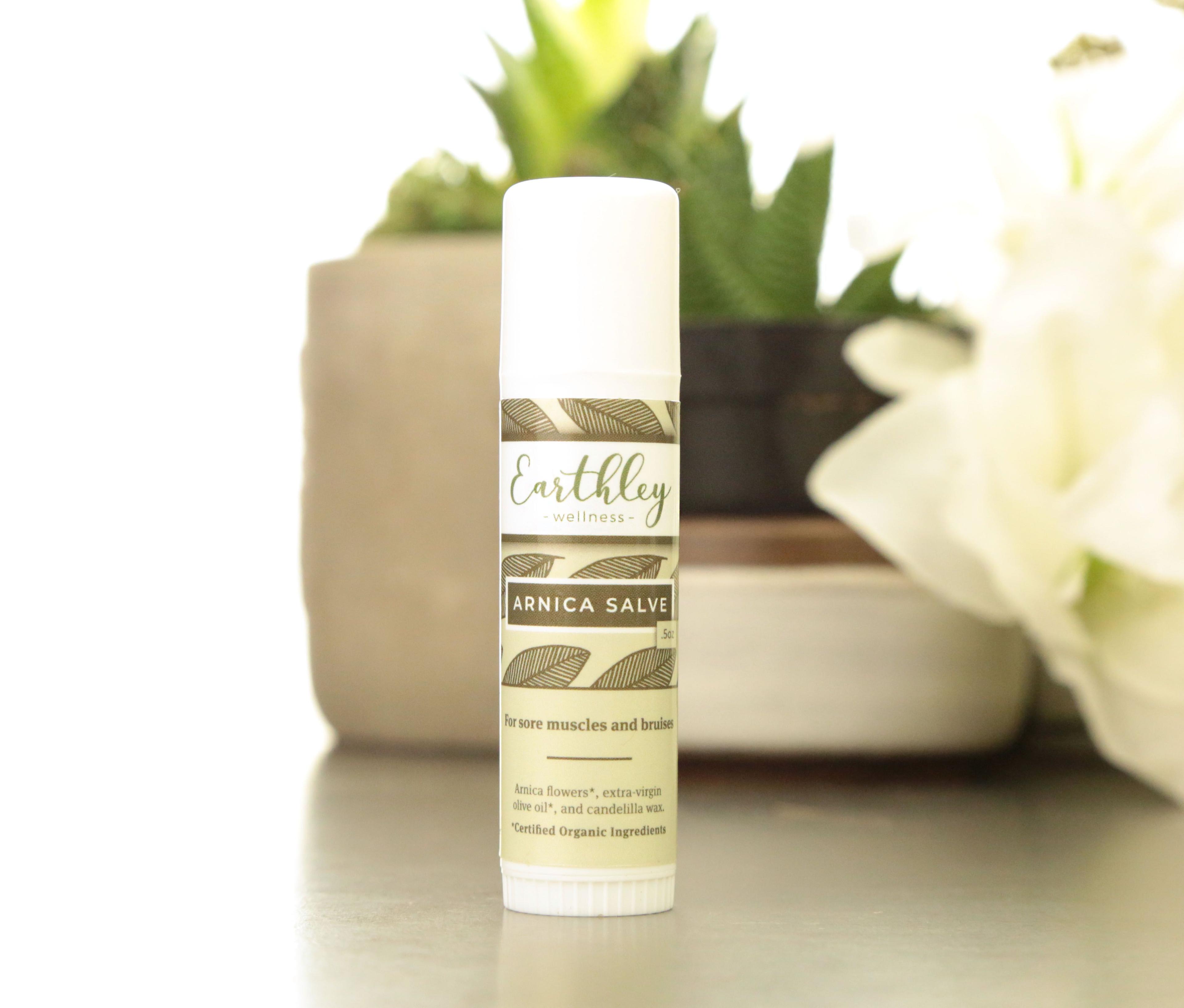 Earthley Arnica Salve twist-up tube with cream and green leaf pattern label showing For sore muscles and bruises text with succulent and white flowers in background
