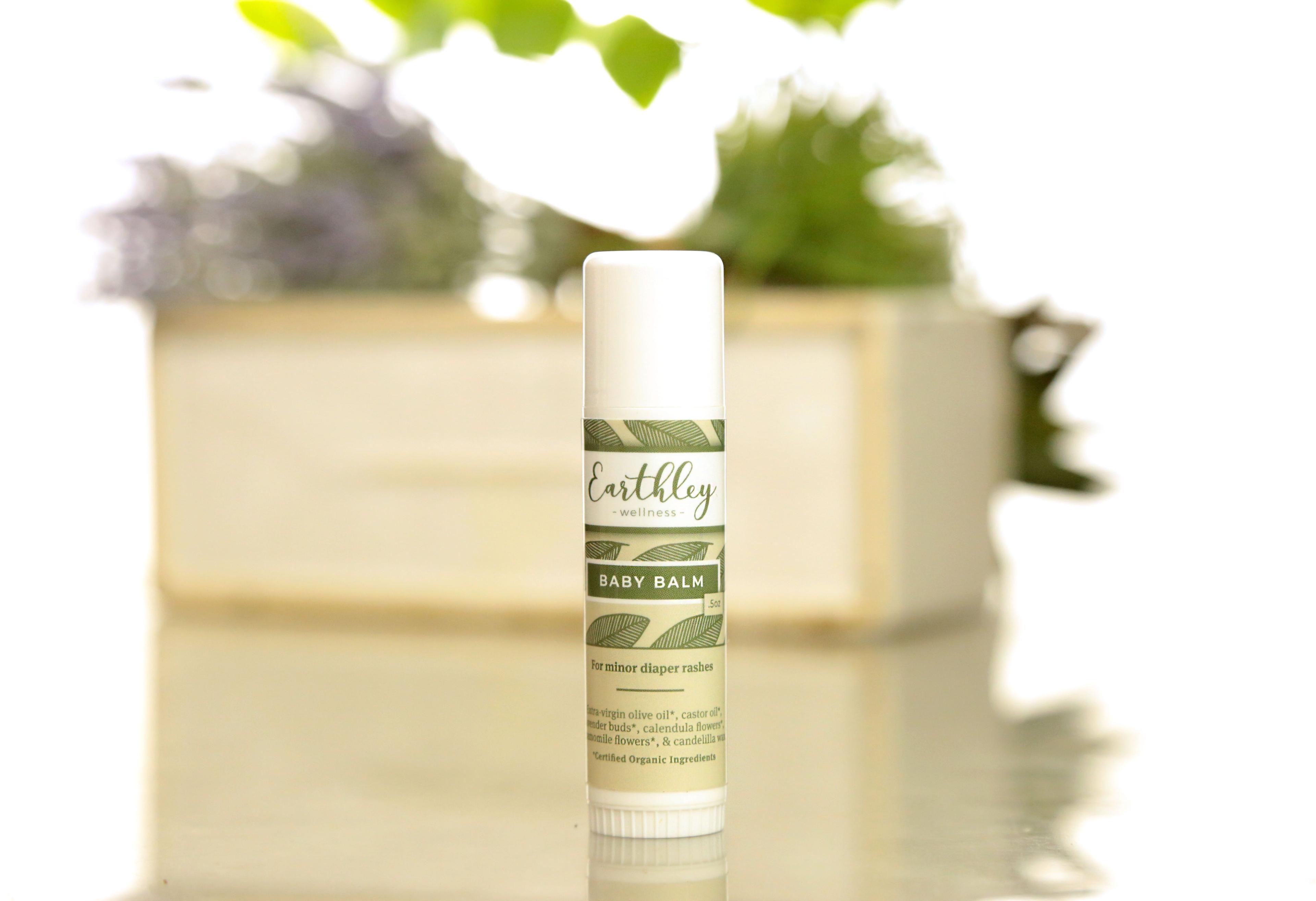 Earthley Baby Balm twist-up tube with green leaf label showing For minor diaper rashes with potted plants and lavender in background on reflective surface