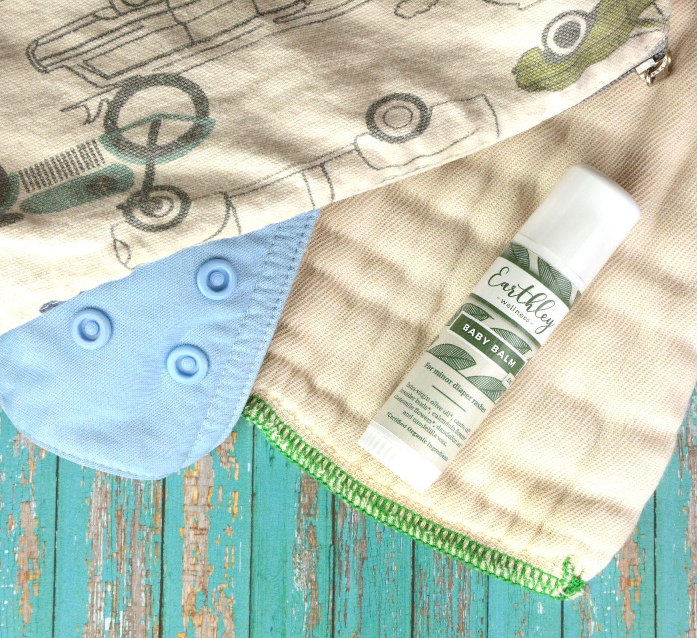 Earthley Baby Balm 0.5oz white frosted twist-up stick with sage green leaf pattern label on tan cream striped baby blanket with blue cloth diaper cover with navy blue snaps on teal turquoise wooden surface