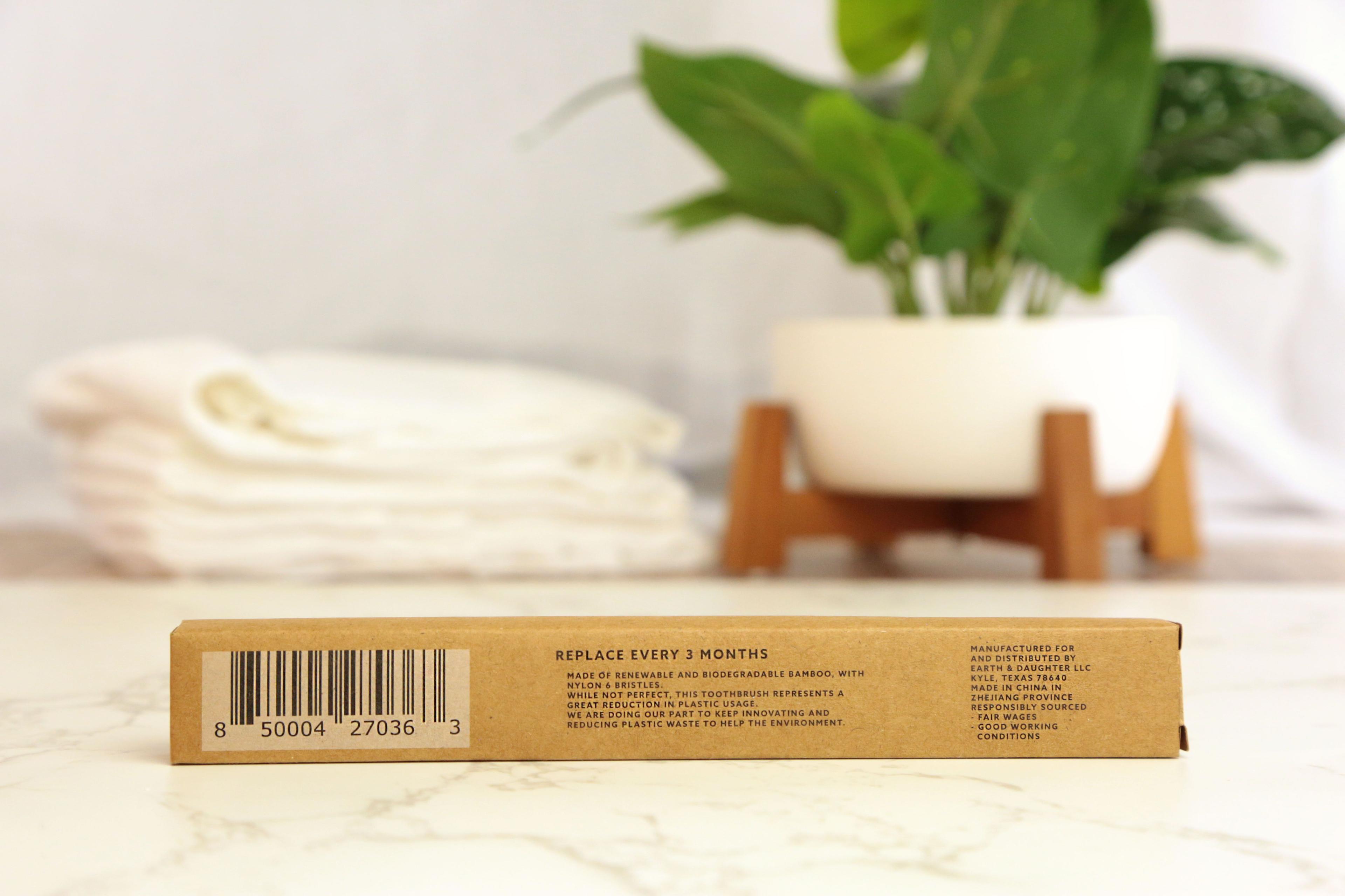 Bamboo toothbrush packaging back view showing tan kraft cardboard box with barcode on white marble with cream towels and green plant in white pot on wooden stand