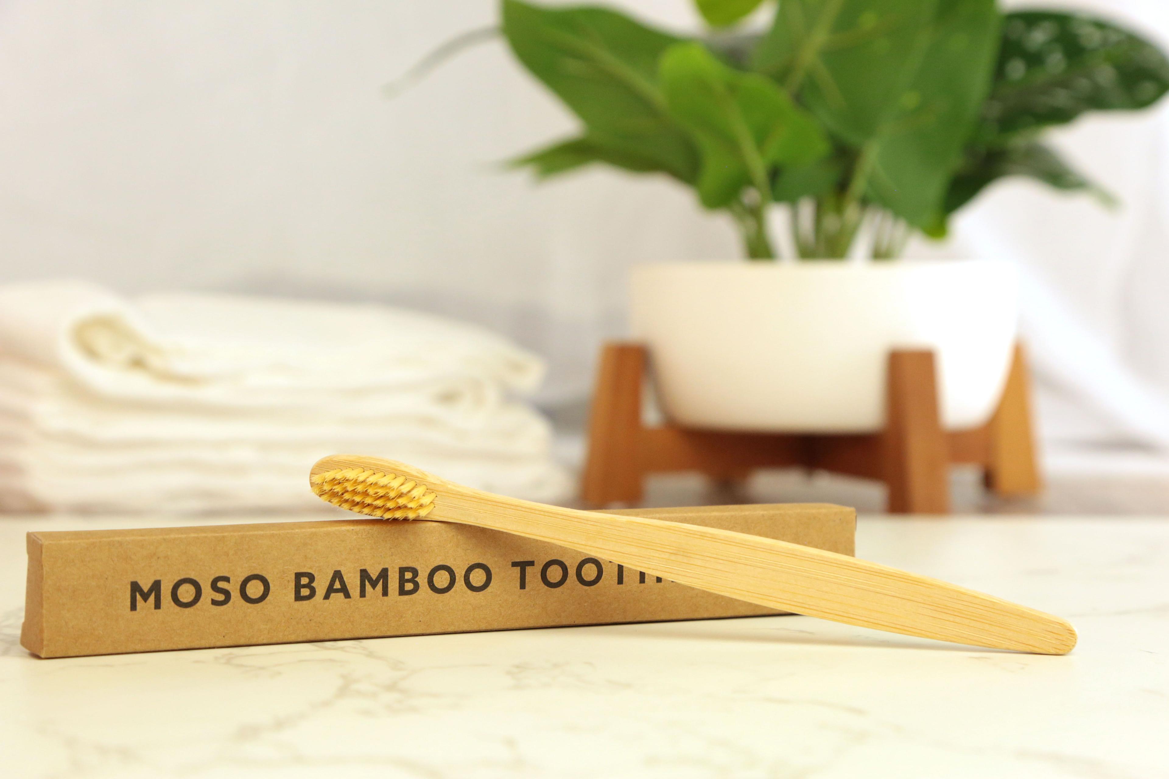 Moso Bamboo Toothbrush with natural tan handle and gold bristles on tan kraft box on white marble with cream towels and green plant in white pot on wooden stand