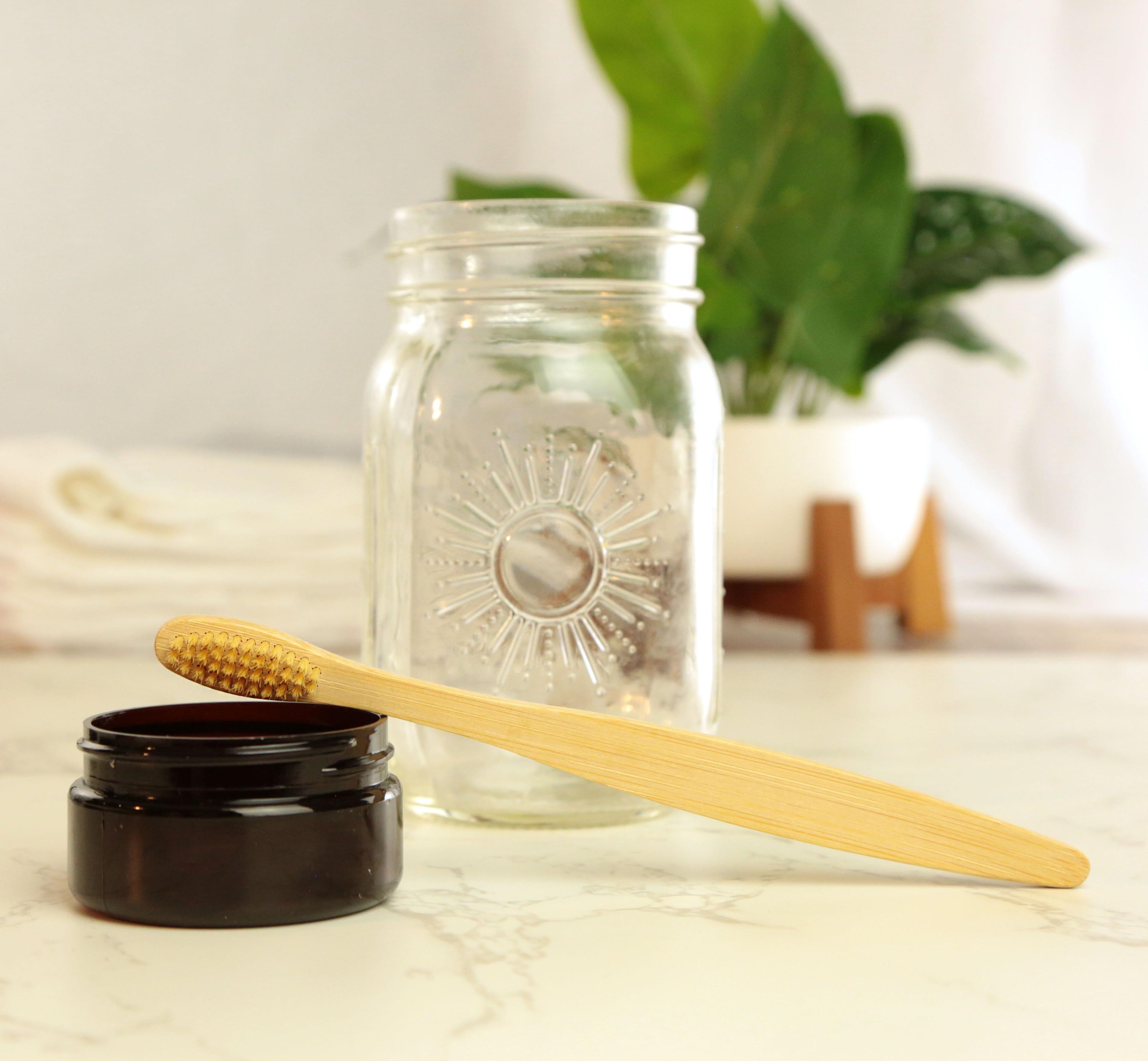 Bamboo toothbrush with natural tan handle and gold bristles resting on amber glass jar on white marble with clear mason jar and green plant in white pot on wooden stand