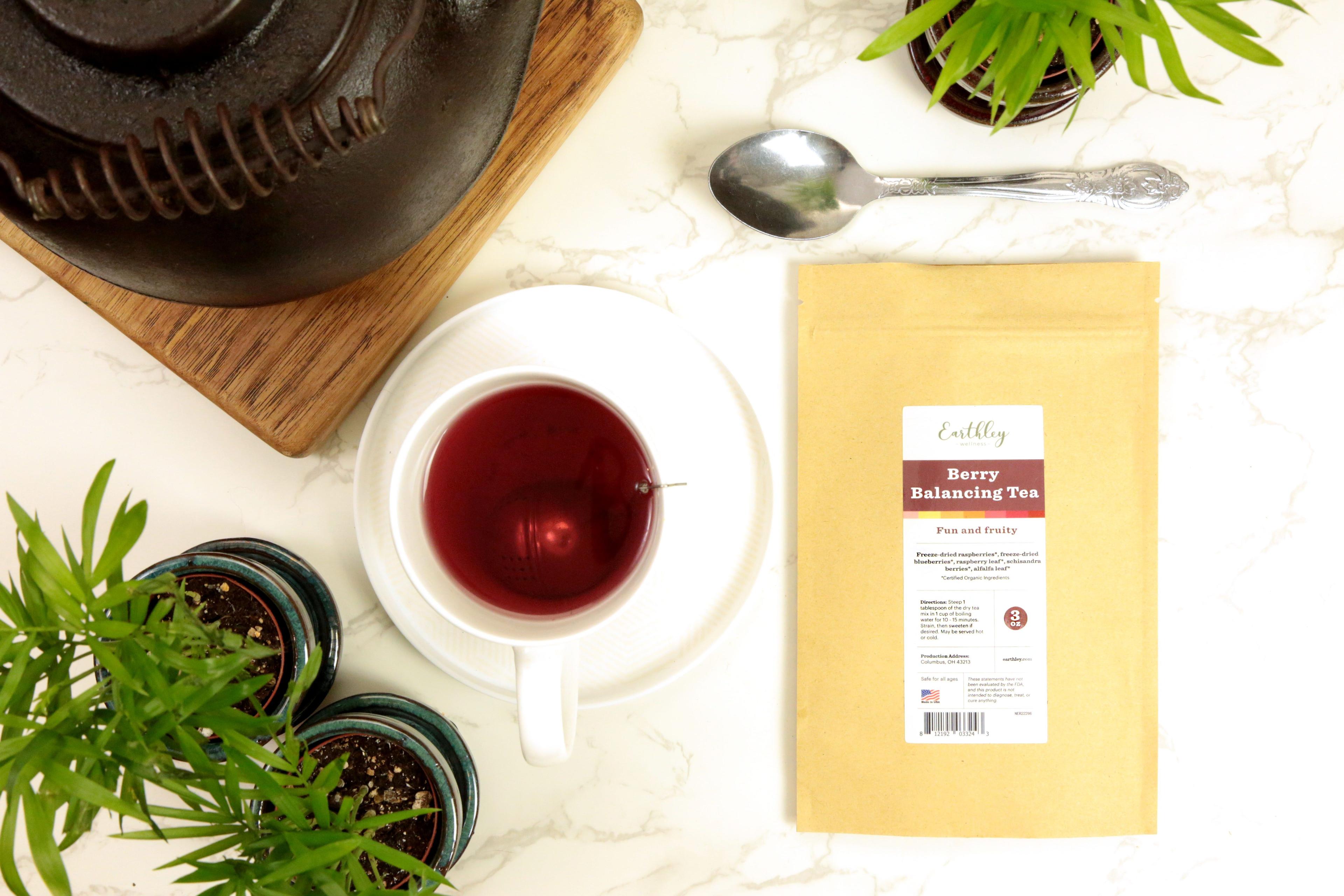 Earthley Berry Balancing Tea 3oz tan kraft resealable pouch with burgundy red and yellow label on white marble with dark brown cast iron teapot wooden board white cup with dark red berry tea silver spoon and green plants in teal pots