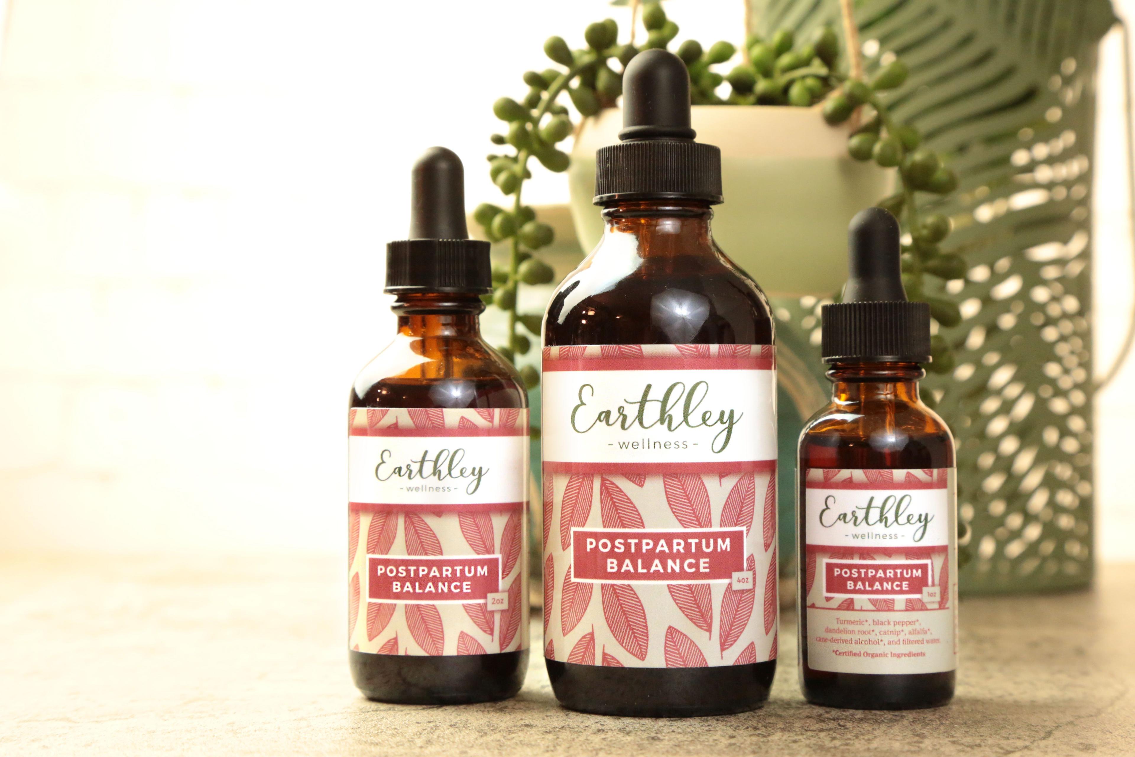 Earthley Postpartum Balance amber glass dropper bottles in three sizes 1oz 4oz and 2oz with black caps and coral pink leaf pattern labels on tan surface with green succulent and fern plants in background