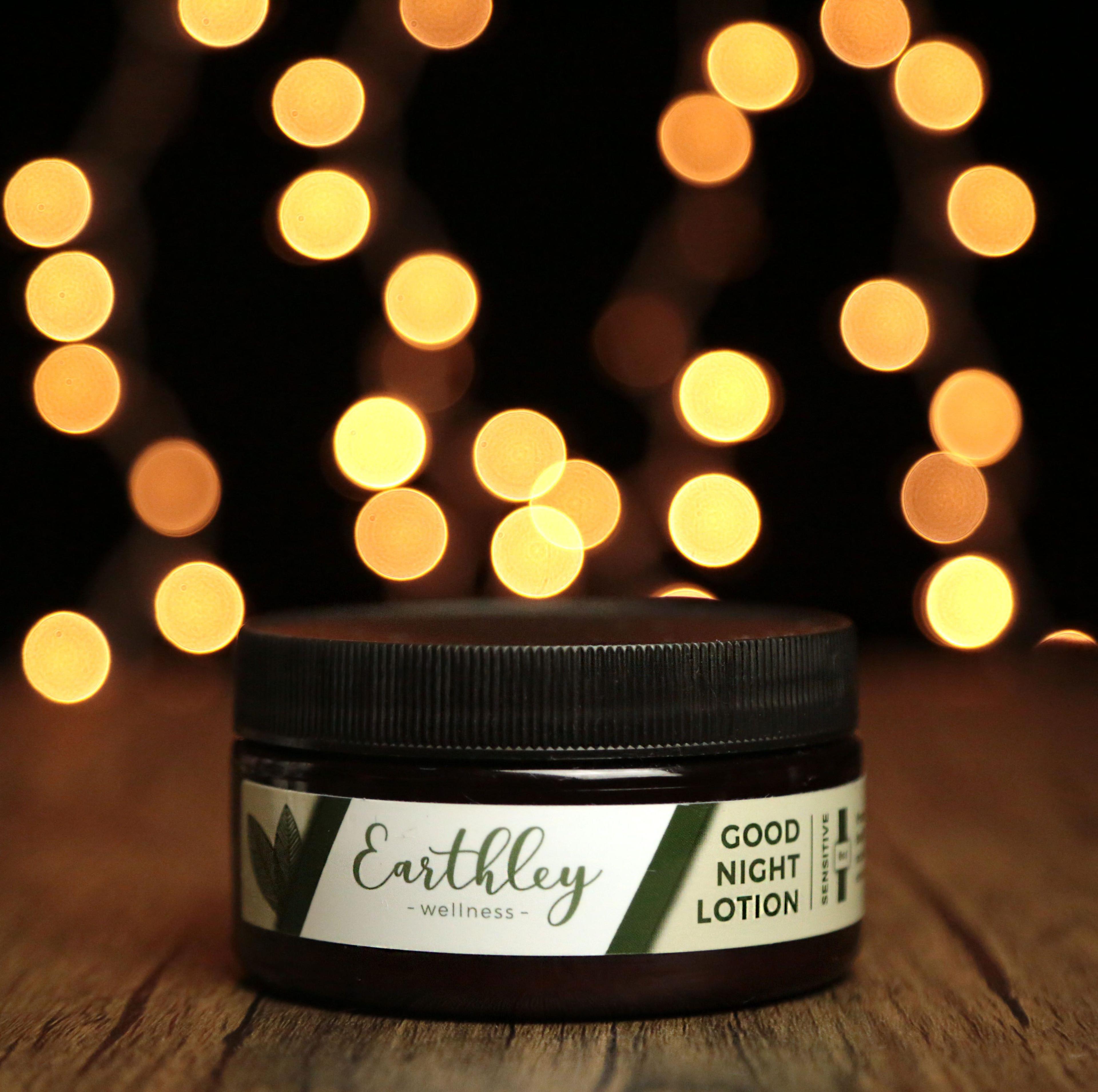 Earthley Good Night Lotion Sensitive amber jar with black ribbed lid and green leaf label on rustic wooden surface with warm golden bokeh lights against dark background
