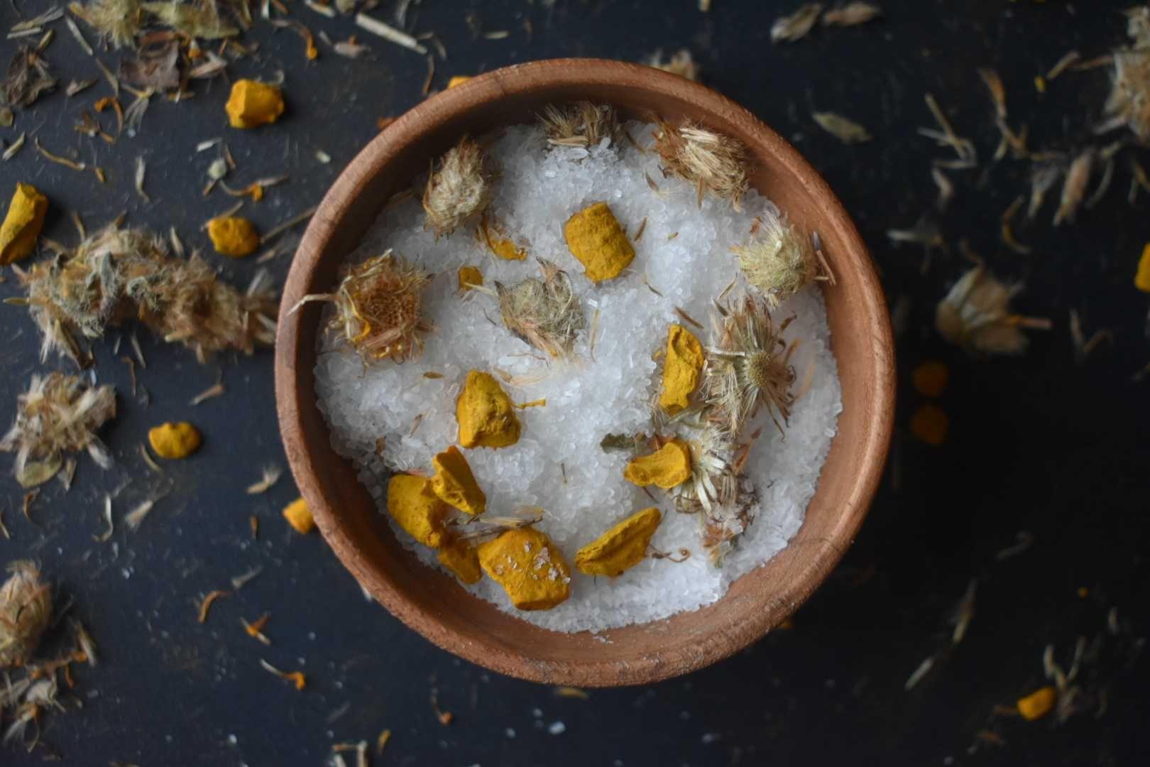 Wooden bowl filled with sea salt mixed with dried chamomile flowers and yellow turmeric pieces on dark surface