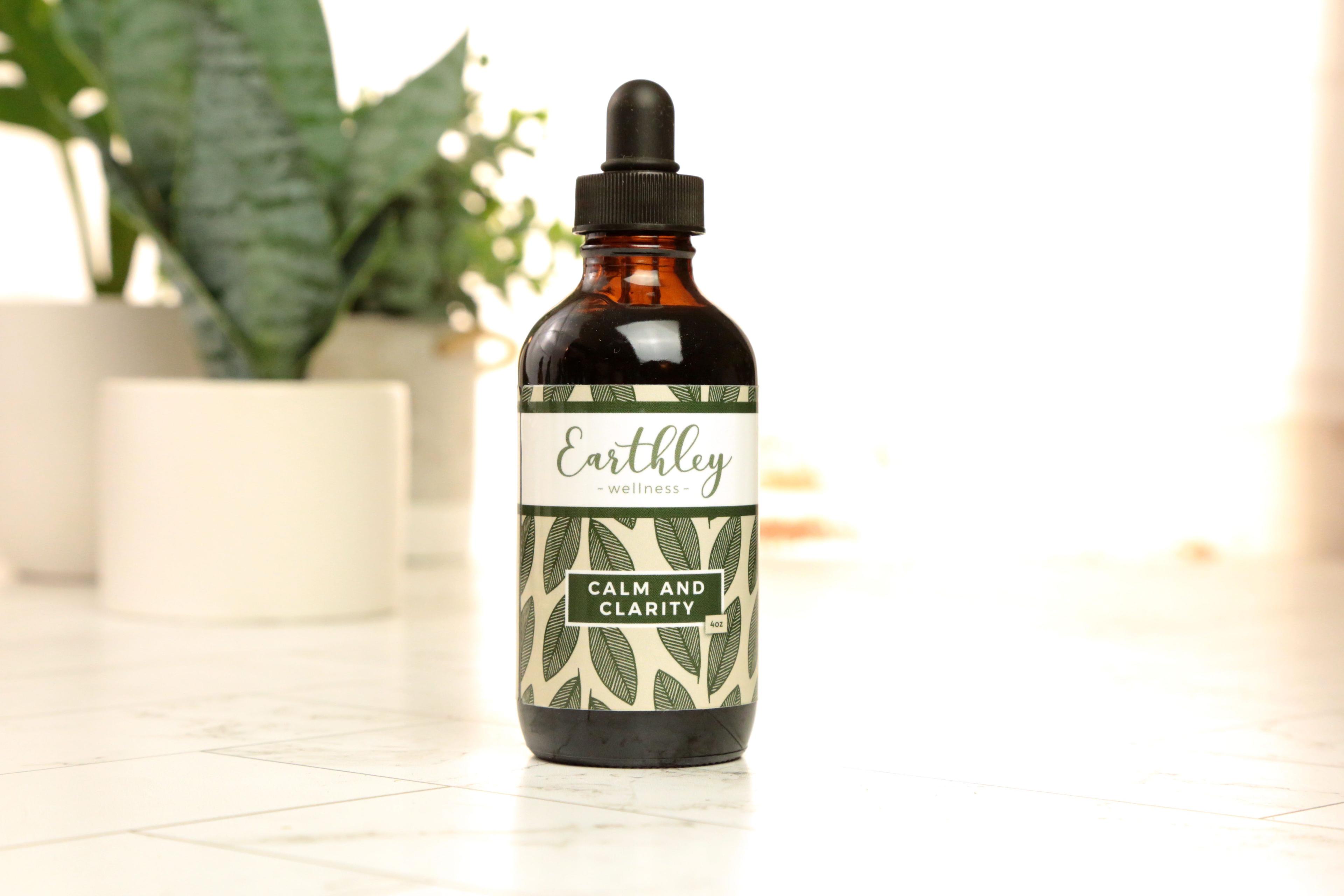 Earthley Calm and Clarity 4oz amber glass dropper bottle with black cap and sage green leaf pattern label on white marble tile with green plants in white ceramic pots