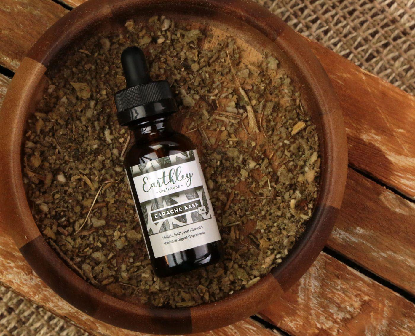 Earthley Earache Ease amber dropper bottle with teal geometric label resting in wooden bowl of dried herbs on wooden surface