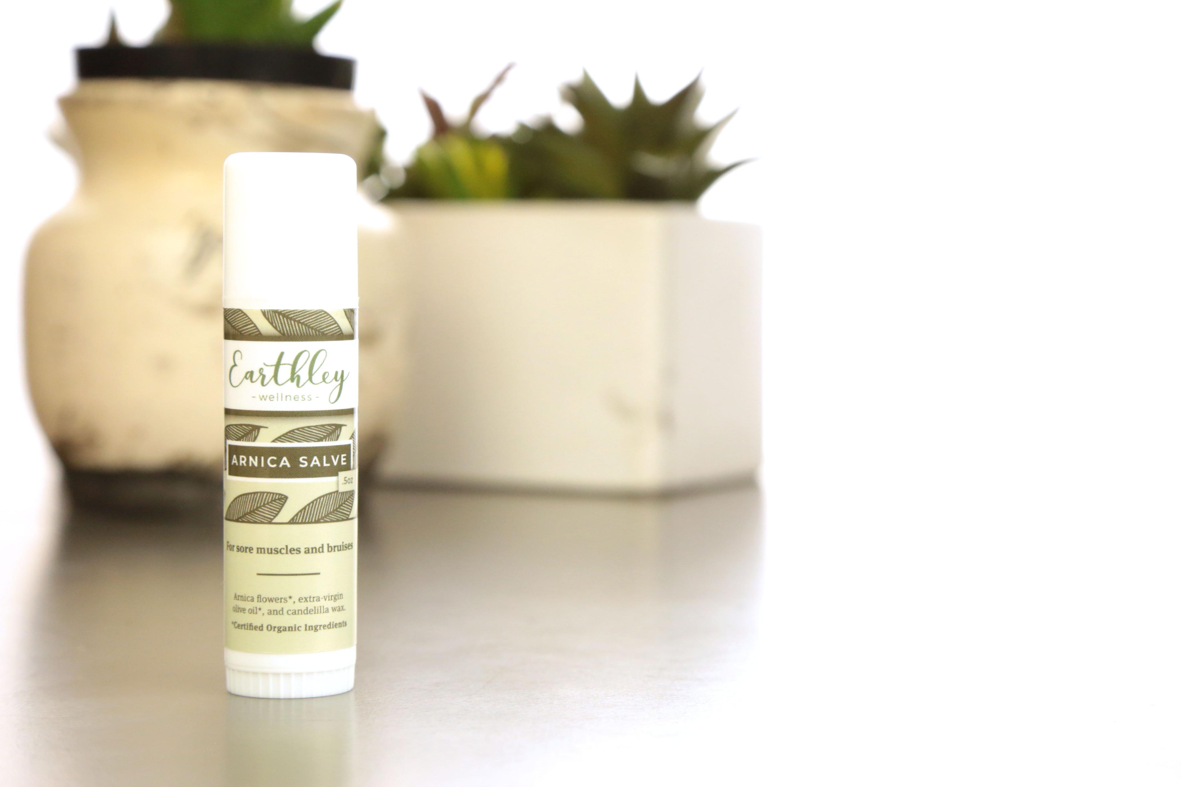 Earthley Arnica Salve white twist-up tube with white cap and sage green leaf pattern label on white surface with green succulents including dark red hens and chicks in cream white and geometric angular ceramic pots soft focus background