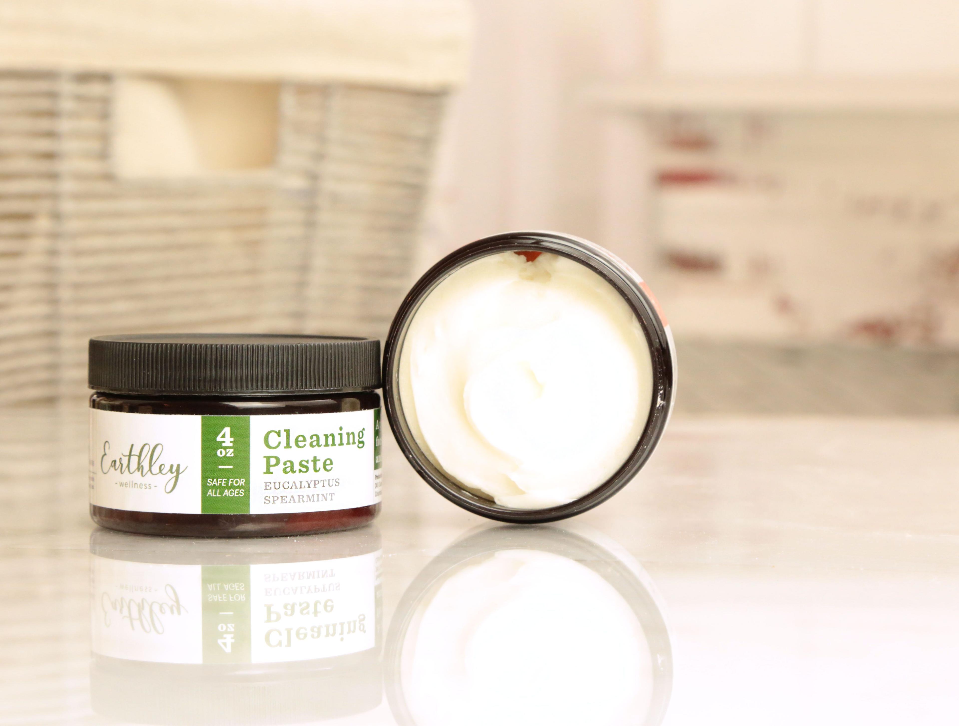 Earthley Cleaning Paste 4oz amber jar with white creamy paste inside eucalyptus spearmint scent safe for all ages for natural household cleaning on reflective surface with wicker basket in background