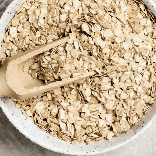 Colloidal oatmeal ingredient photo showing white ceramic bowl filled with rolled oats and wooden spoon on light tan background