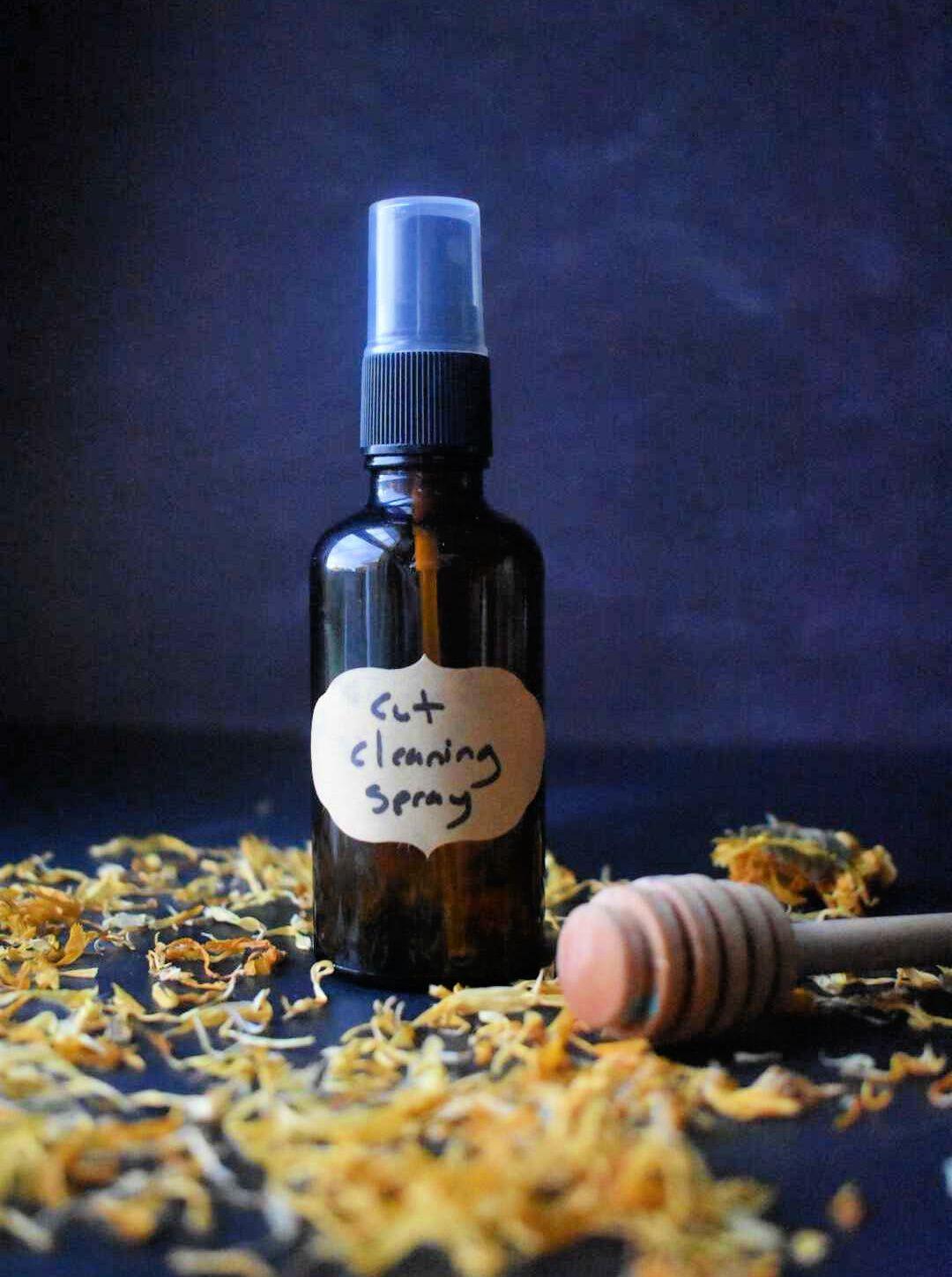 Amber glass spray bottle labeled "Cut Cleaning Spray" with wooden honey dipper surrounded by dried calendula petals