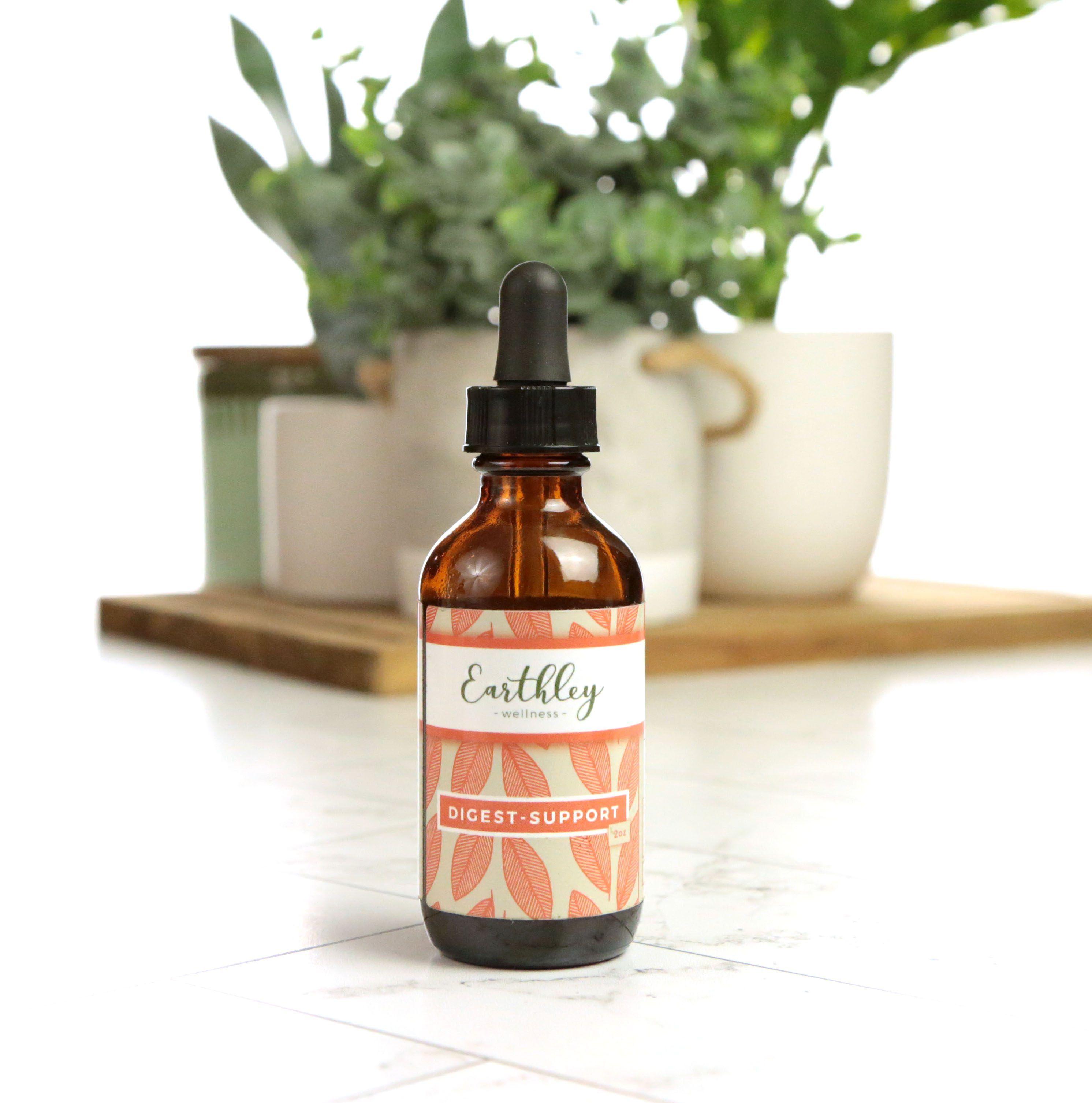 Earthley Digest Support 2oz amber glass dropper bottle with black cap and coral orange leaf pattern label on white marble tile with green plant in cream mug on wooden cutting board