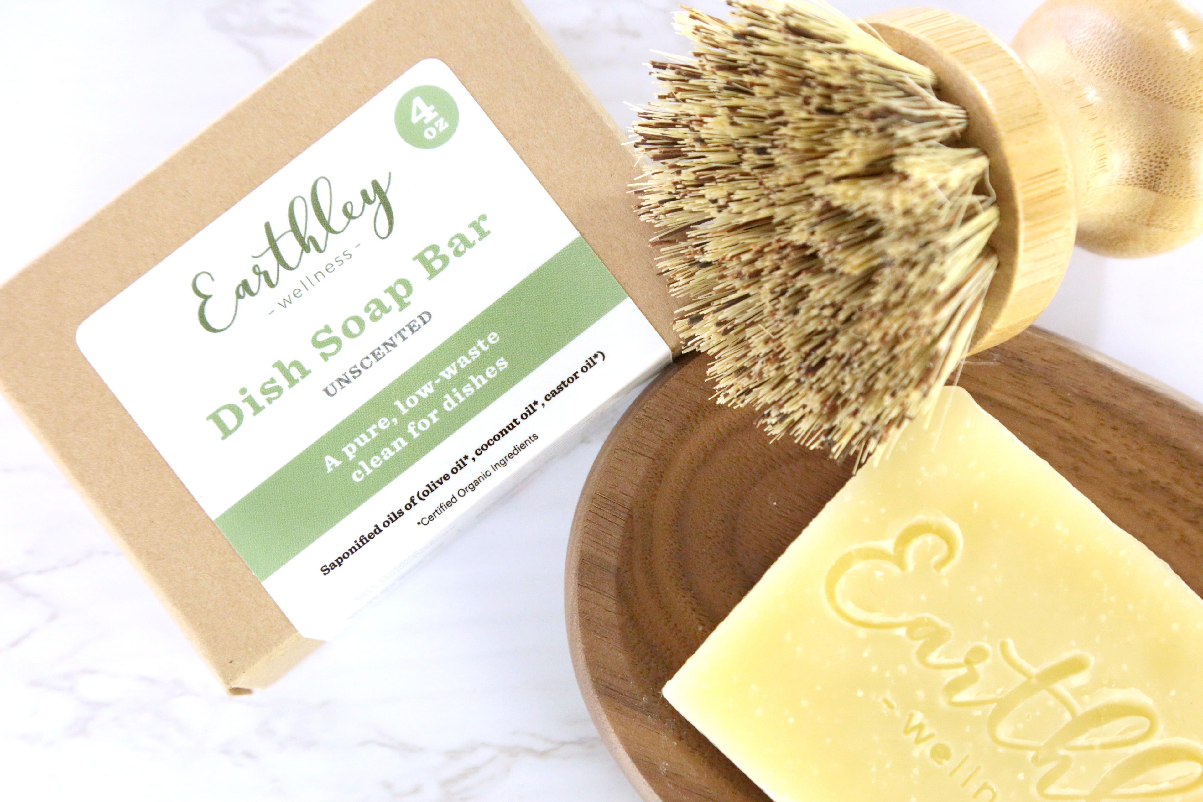 Earthley Dish Soap Bar Unscented 4oz tan kraft box with green stripe label and pale yellow soap bar with Earthley logo embossed on dark brown wooden dish with tan bristle brush on white marble