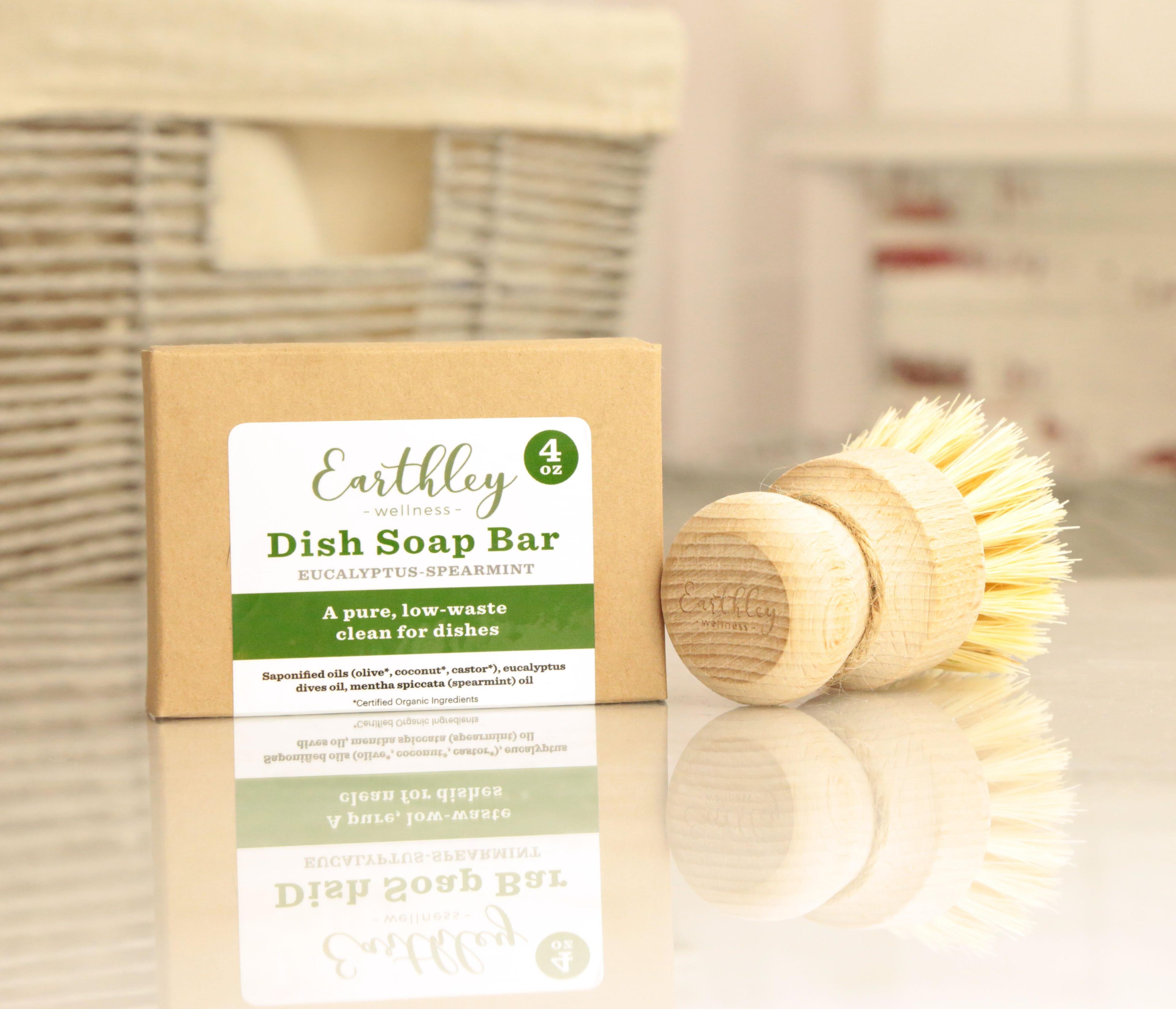 Earthley Dish Soap Bar Eucalyptus Spearmint 4oz tan kraft box with green stripe label and cream wooden bristle brush on reflective white surface with tan woven basket in background