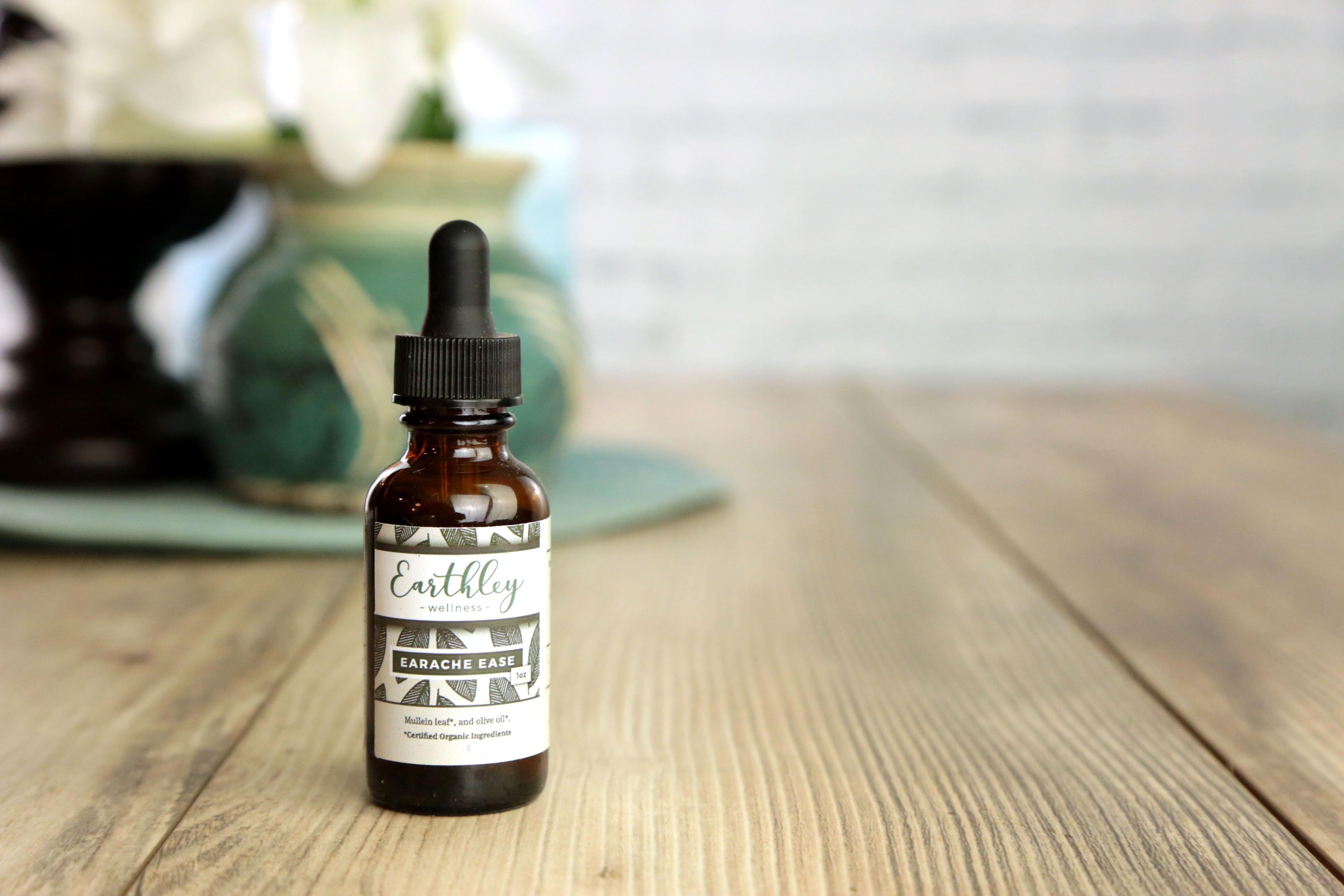 Earthley Earache Ease ear oil bottle with geometric pattern label on wooden surface with teal vase and white flowers