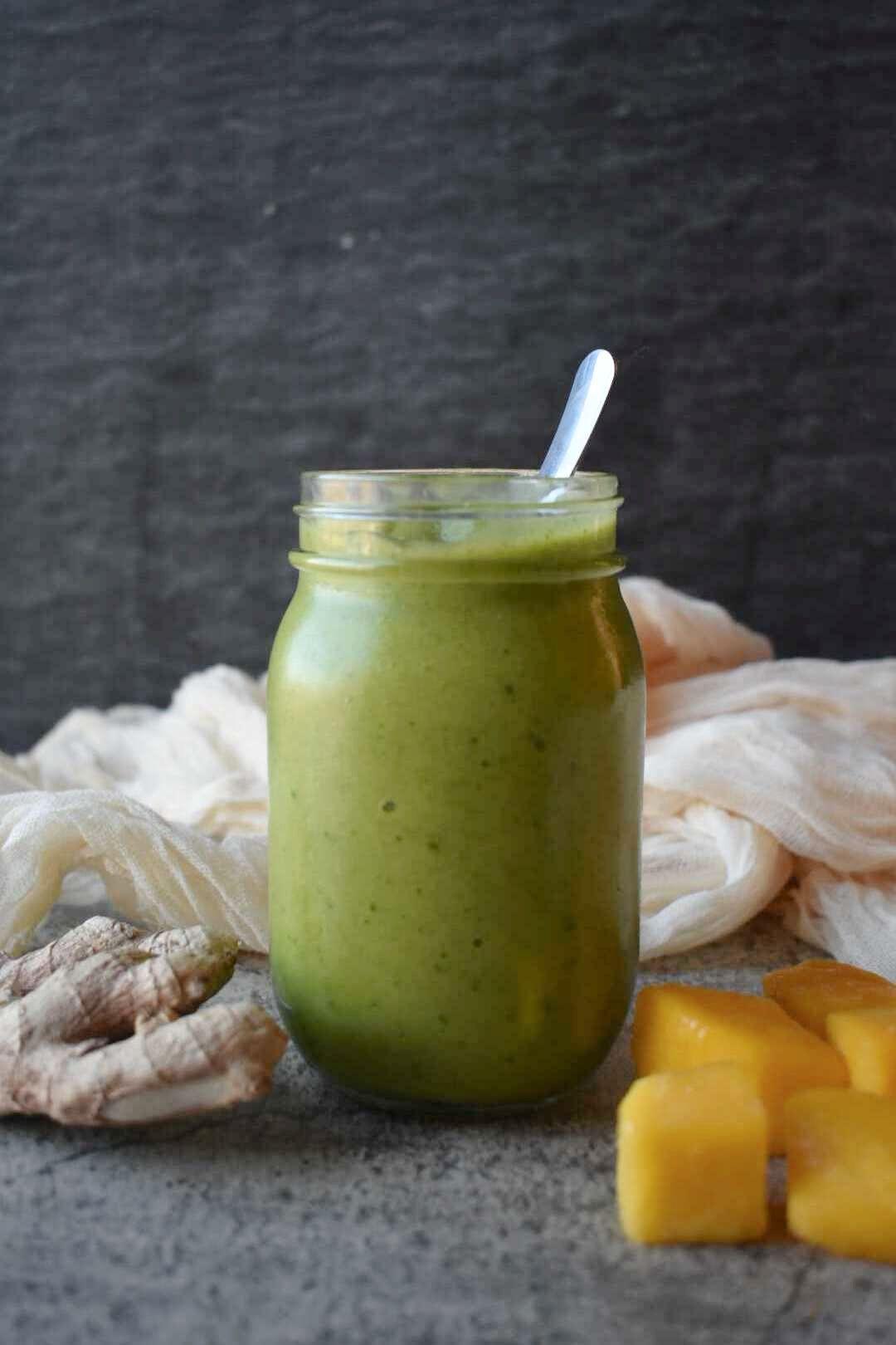 Green smoothie in mason jar with white straw, surrounded by fresh ginger root and mango chunks on gray surface