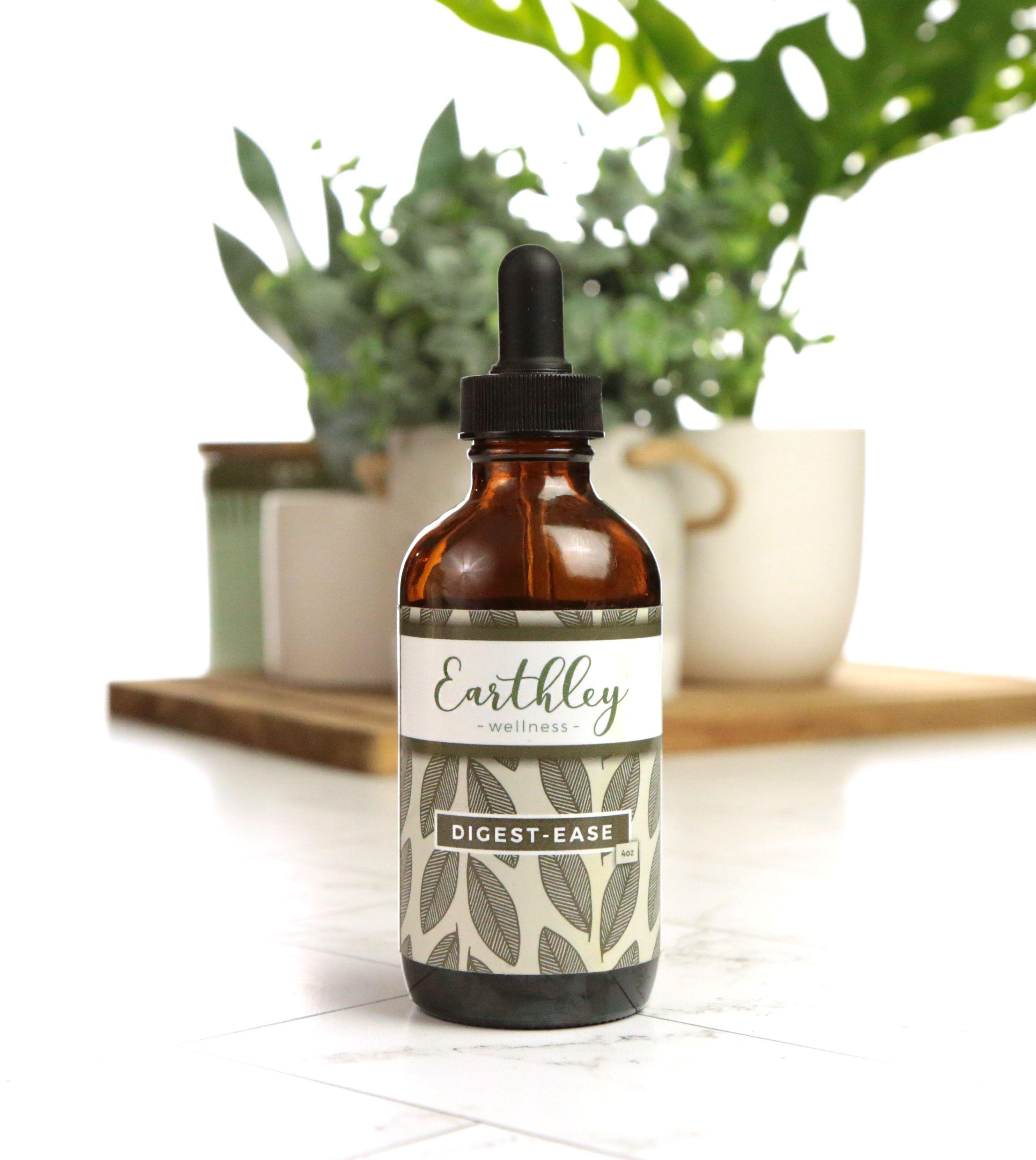 Earthley Digest-Ease 4oz amber glass dropper bottle with black cap and sage green leaf pattern label on white marble surface with green monstera and trailing plants in sage green ribbed ceramic pot and cream ceramic mug on wooden cutting board