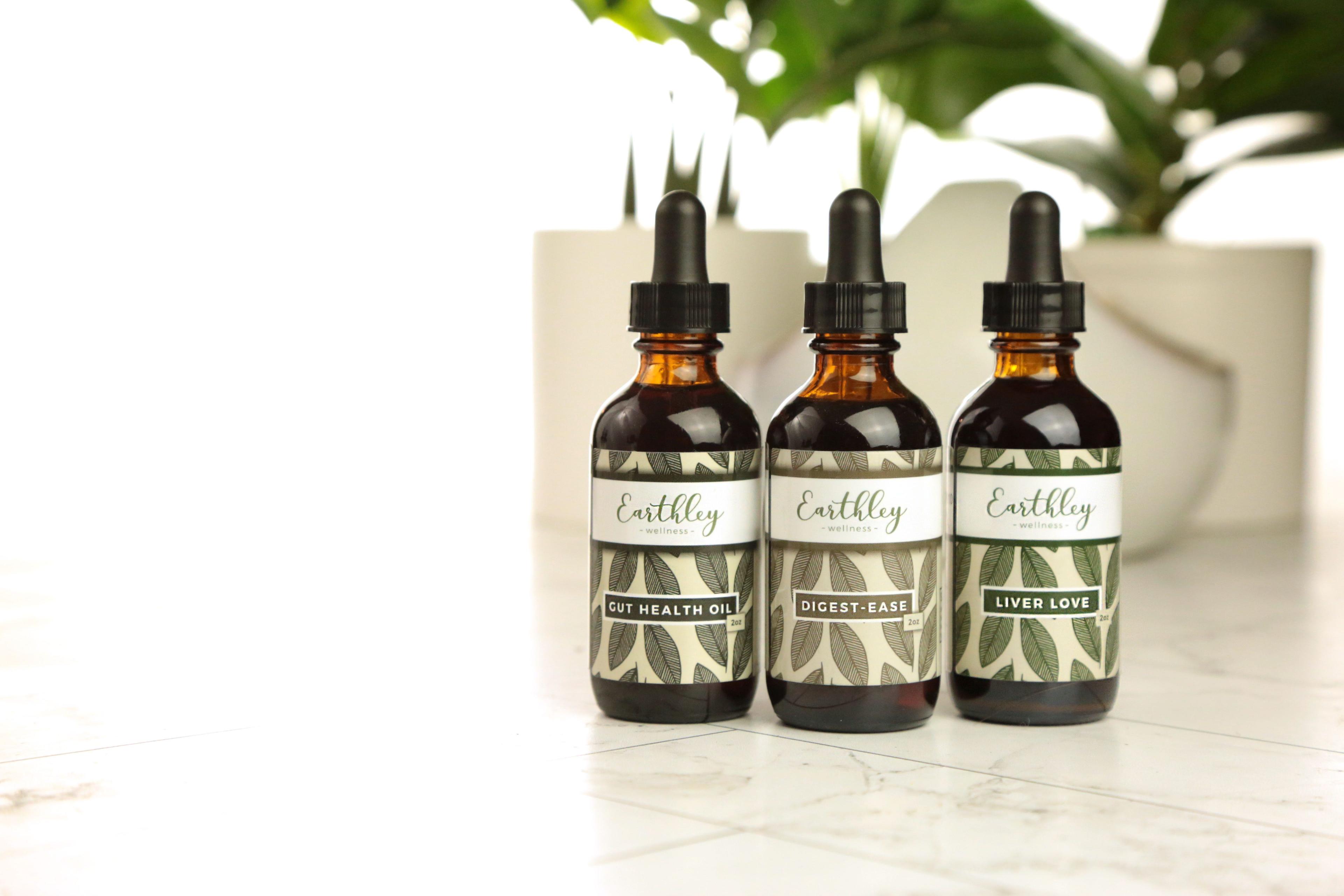 Earthley Gut Health Bundle featuring three amber glass dropper bottles with black caps showing Gut Health Oil Digest-Ease and Liver Love all with sage green leaf pattern labels arranged on white marble surface with green monstera leaves in background