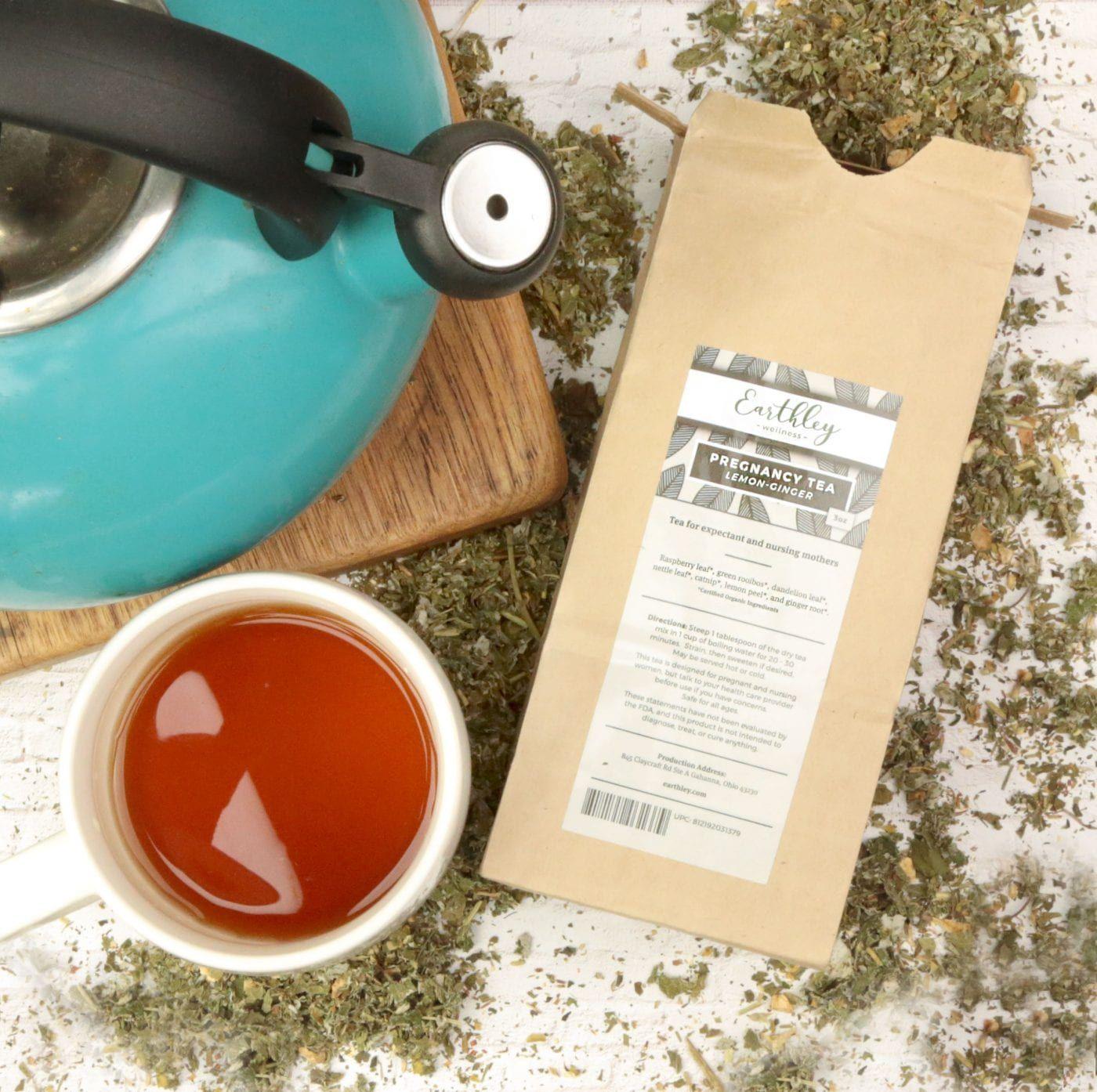 Earthley Pregnancy Tea Lemon Ginger tan kraft resealable pouch with sage green and gray leaf label on white surface with teal aqua kettle wooden cutting board and white cup with amber red tea
