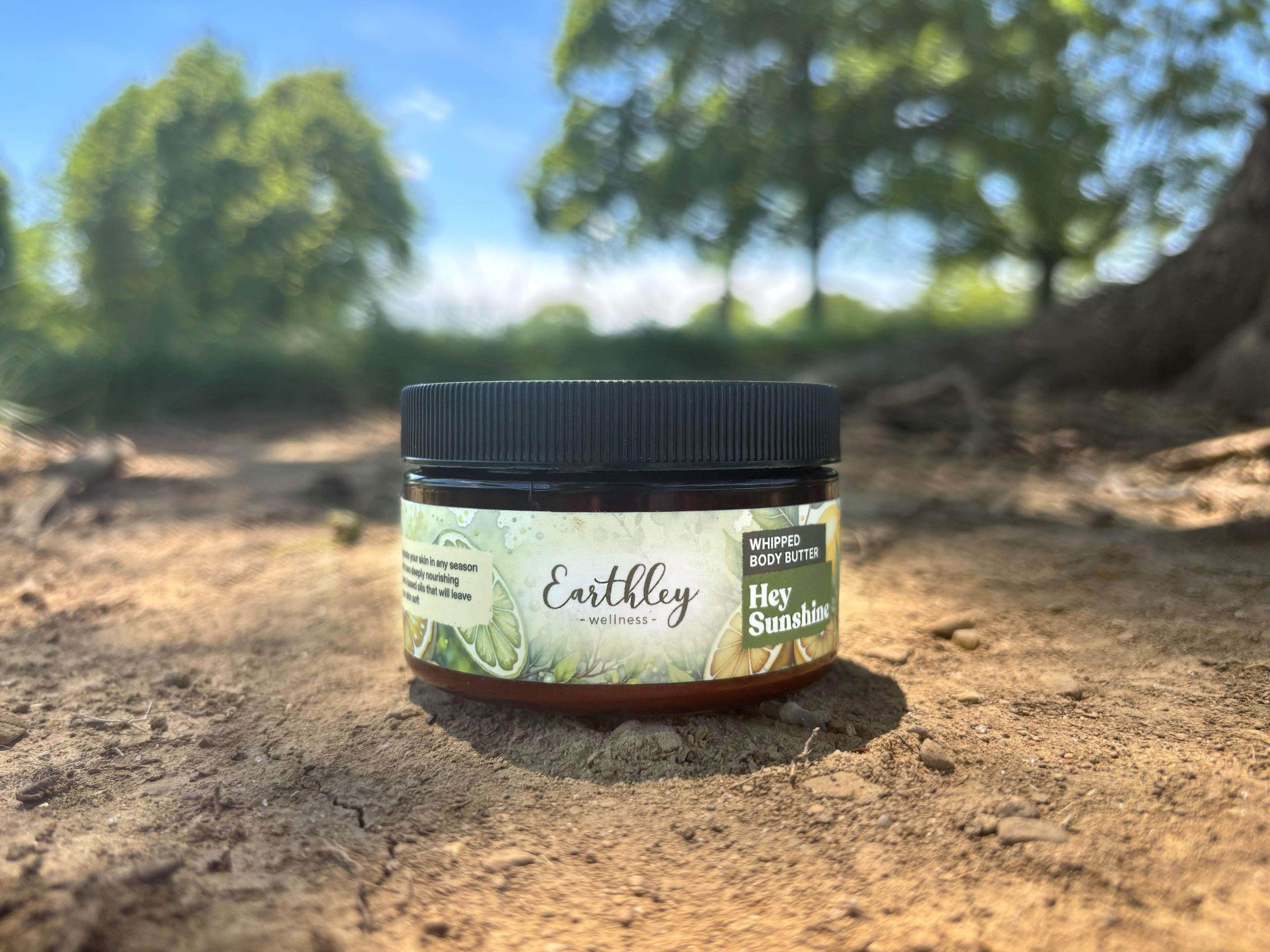Earthley Hey Sunshine Whipped Body Butter 4oz amber jar with black lid and citrus-themed green label photographed outdoors on dry earth with trees and blue sky in background