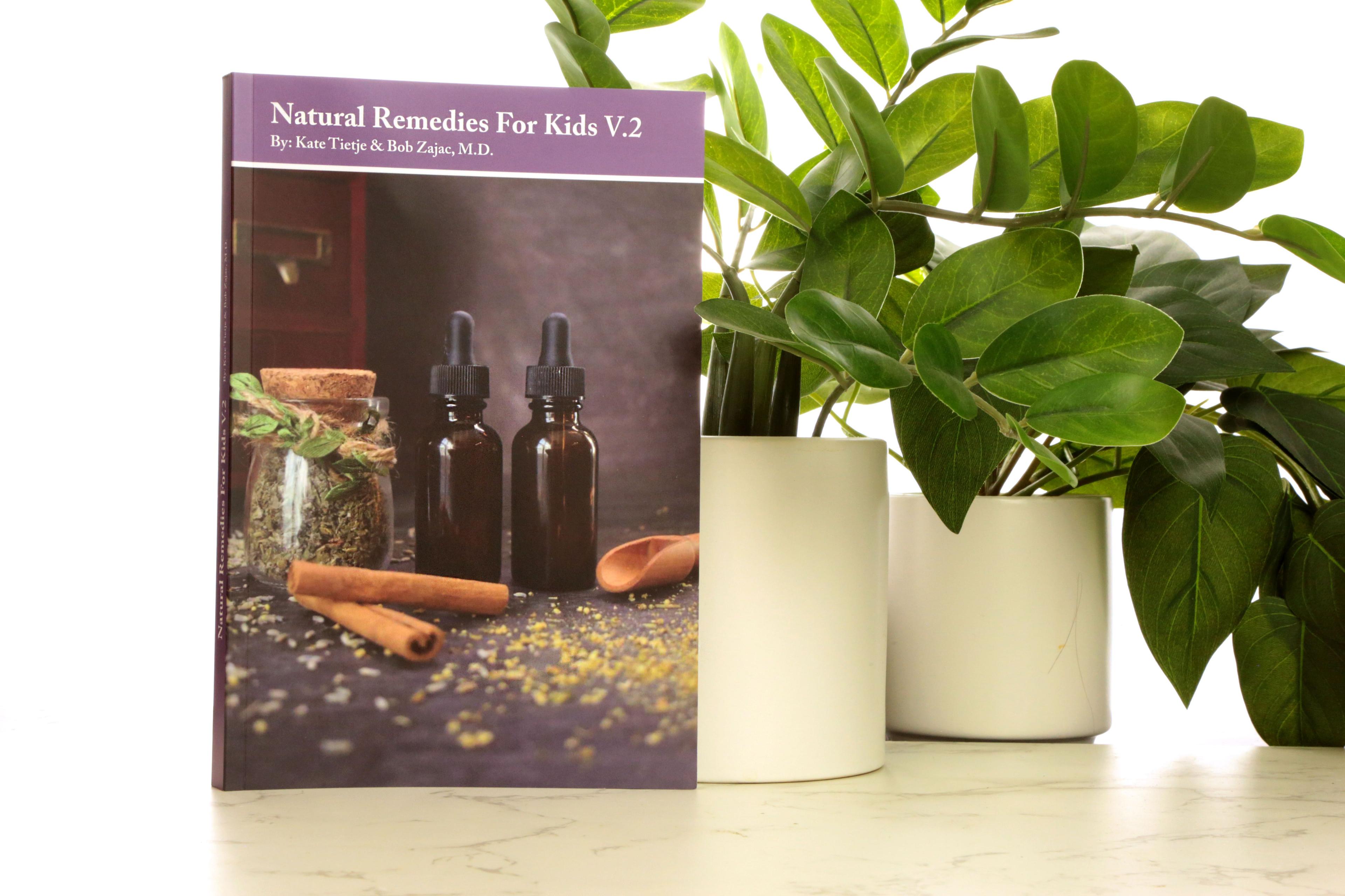 Natural Remedies for Kids V.2 book cover featuring purple header with amber dropper bottles, brown cinnamon sticks, and cork-top glass herb jar on dark gray background, displayed on white marble with green plants in white pots