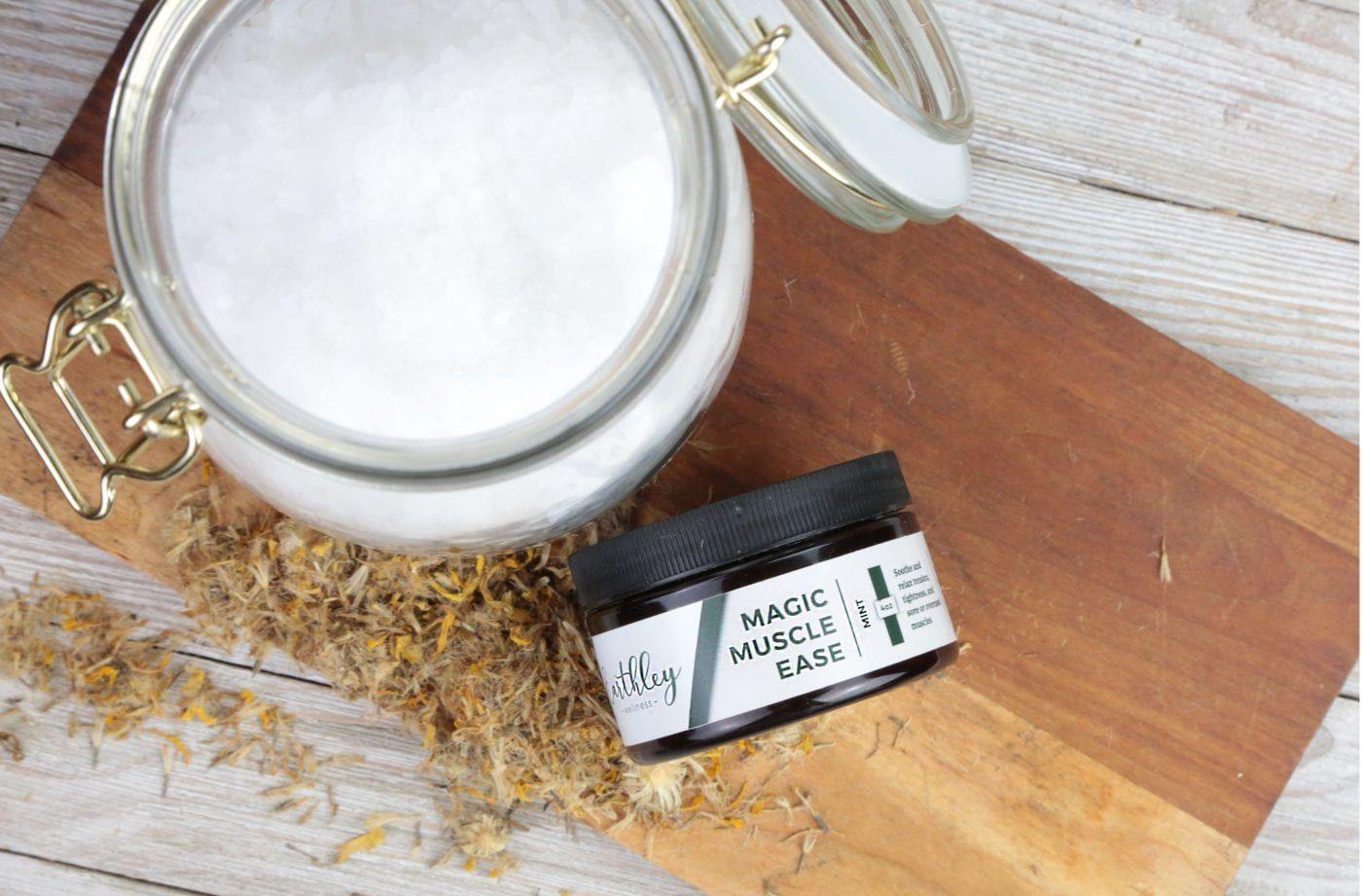 Earthley Magic Muscle Ease jar with green mint label on wooden cutting board with dried calendula flowers and glass jar of salt