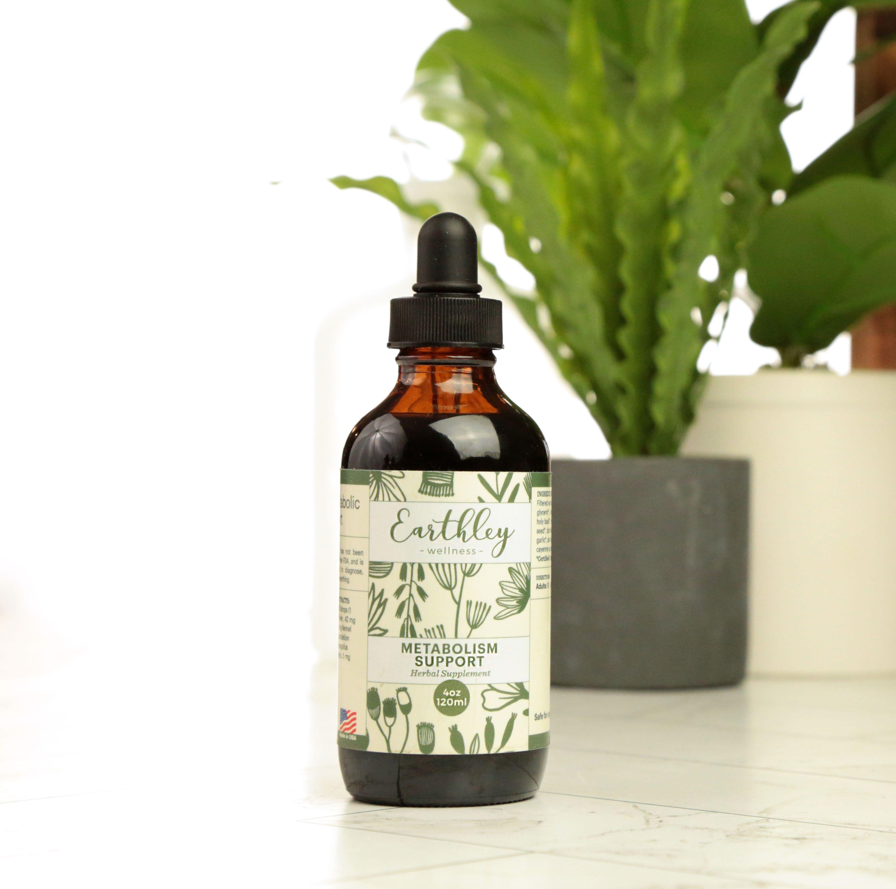 Metabolism Support 4oz product photo showing dark amber glass dropper bottle with black cap and cream label with sage green botanical herb pattern illustrations on white marble surface with bright green fern plant in dark gray pot in background