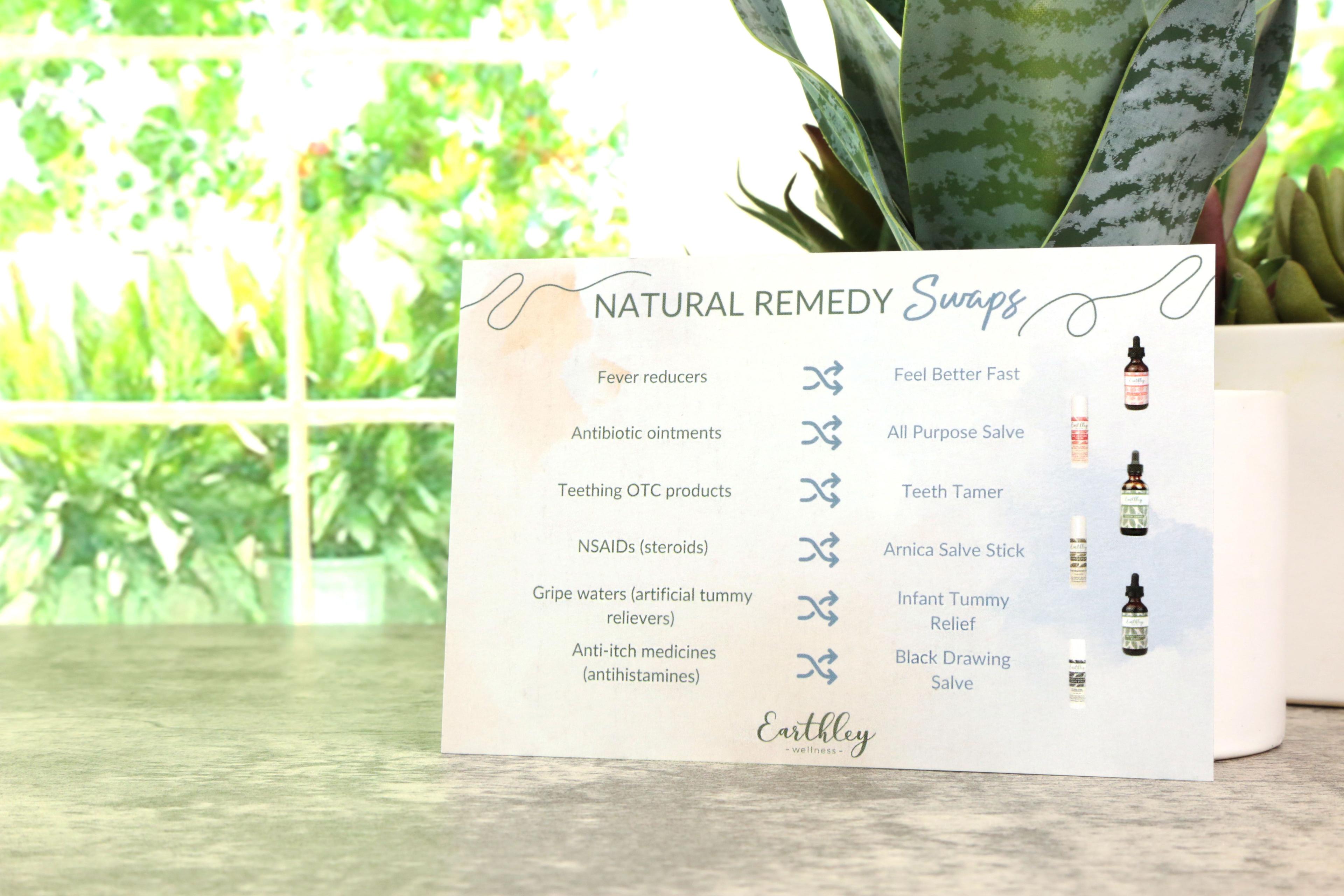 Earthley Natural Remedy Swaps magnet card with teal blue watercolor background showing conventional medicine swaps for Feel Better Fast All Purpose Salve Teeth Tamer Arnica Salve Infant Tummy Relief and Black Drawing Salve with amber bottle images on gray marble surface with green snake plant